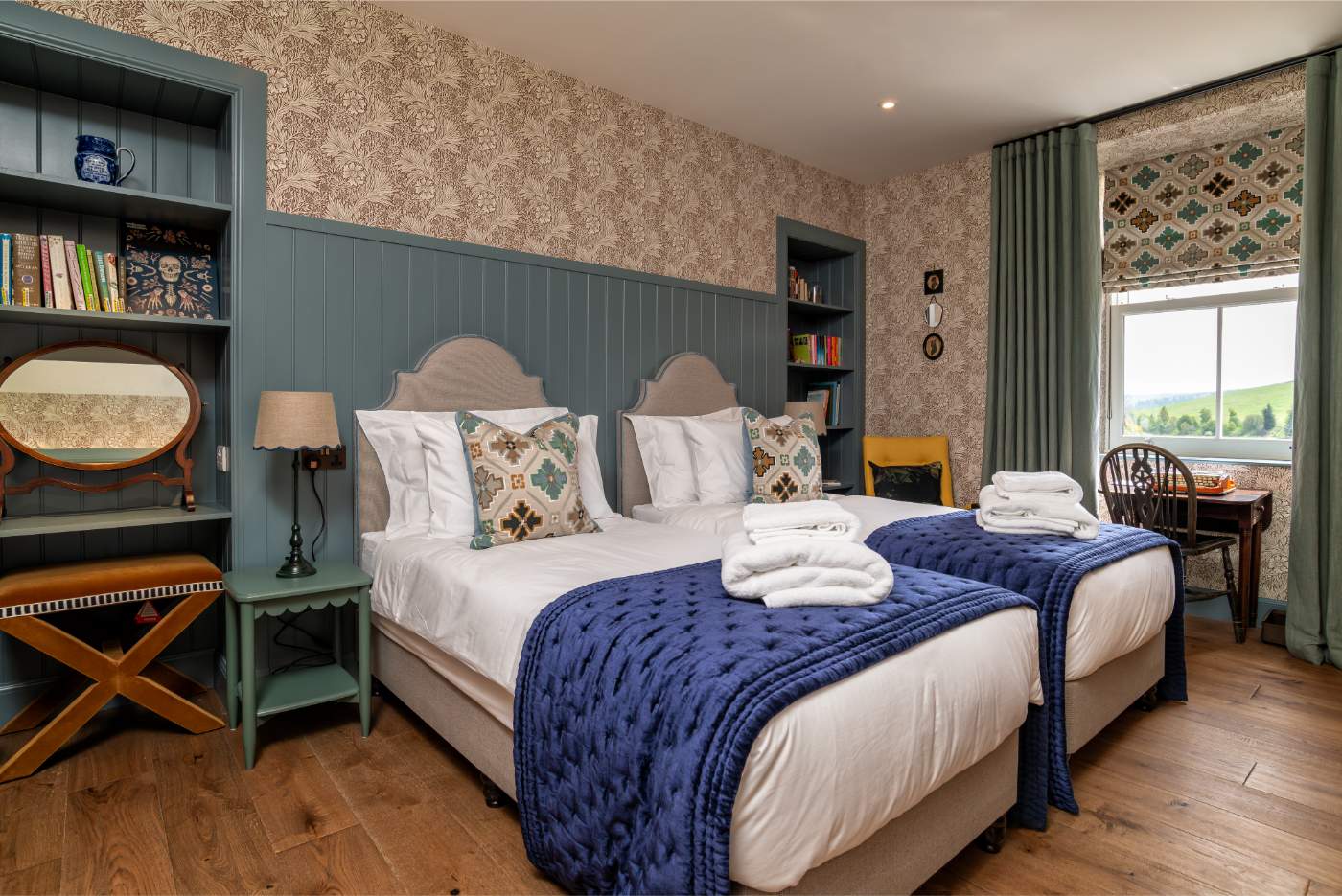 Twin beds with navy throws and folded towels sit beneath a patterned wallpaper, a dressing table and bookshelf in the alcove to the left.