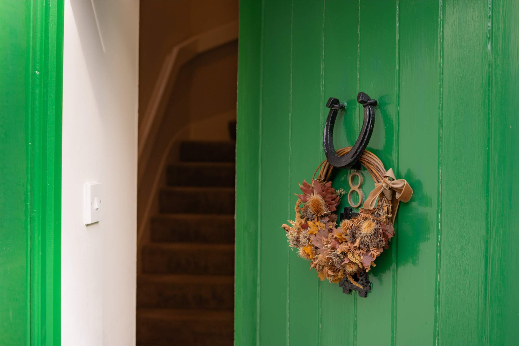 A horseshoe and dried flower wreath with a number hang on a green door, a staircase visible through the open doorway.