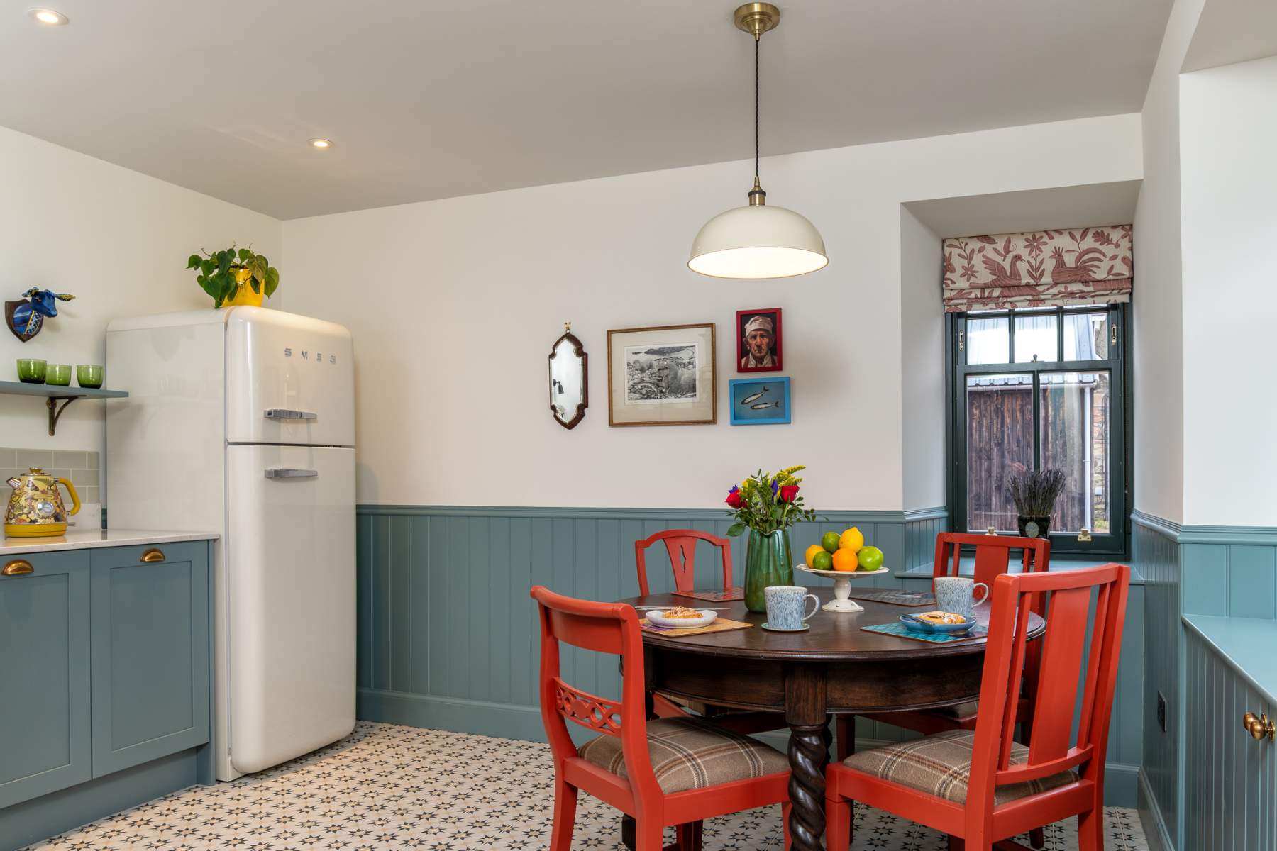 A cream fridge freezer stands beside dusty blue cabinetry, a round table set with fruit in front of a curtained window.