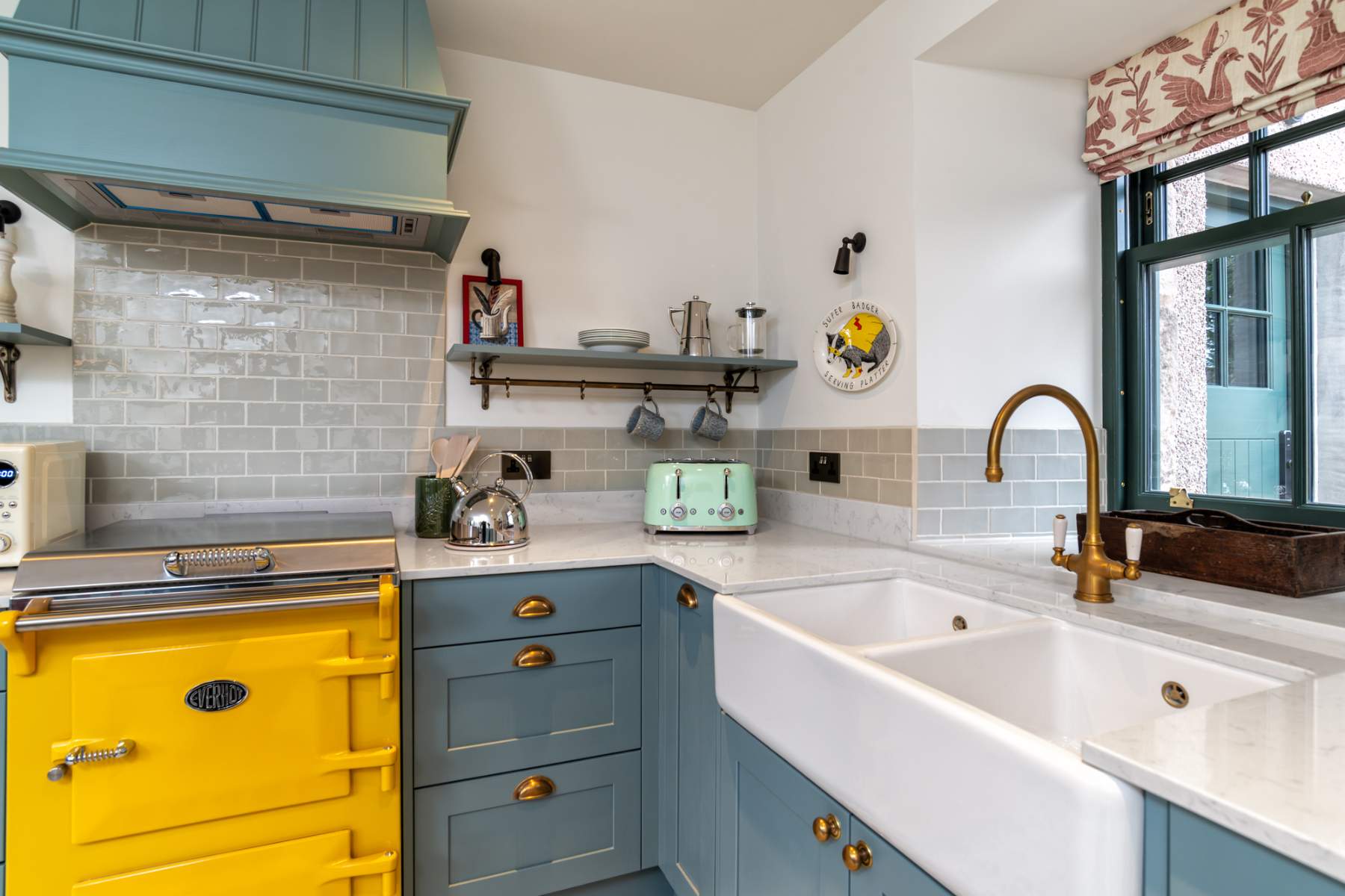 A yellow range cooker sits below an extractor hood in a kitchen with grey tiling, a butler's sink and brass tap beneath a window.