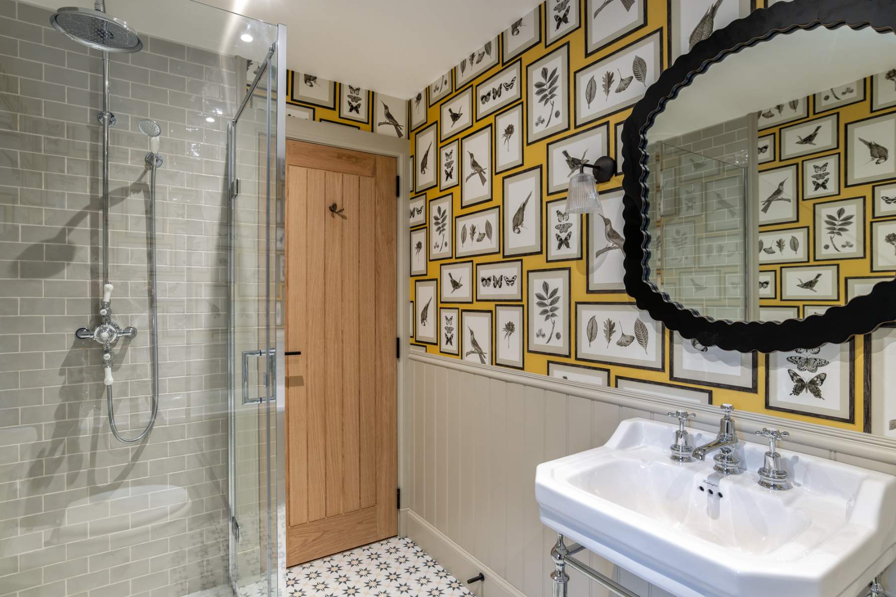 A glass shower cubicle stands beside a pedestal basin and round mirror in a bathroom papered with yellow bird and plant illustrations.