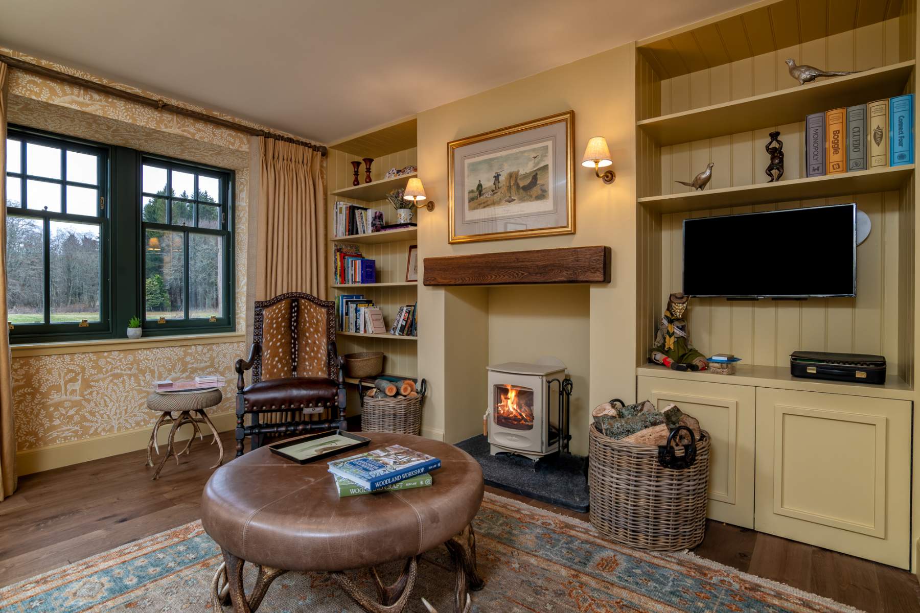 A leather armchair and round footstool sit before a wood burning stove, bookshelves and a mounted television built into the alcoves either side.