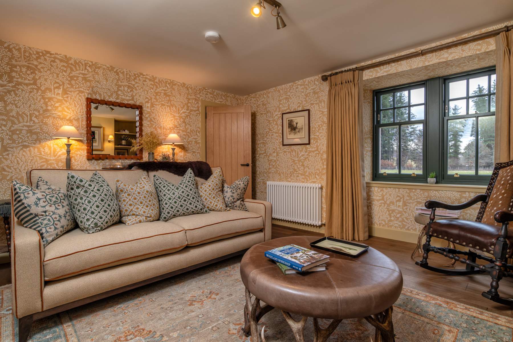 A cream sofa with patterned cushions sits below a gilt mirror, a rocking chair and round leather footstool in the foreground of a patterned-wallpaper sitting room.