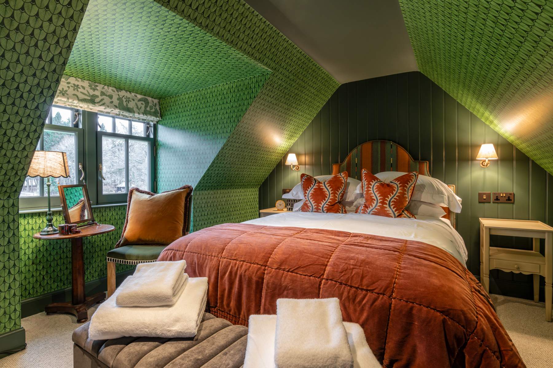 A bed with a rust-coloured throw and patterned cushions sits in a green-papered attic room, folded towels and a stool at its foot.