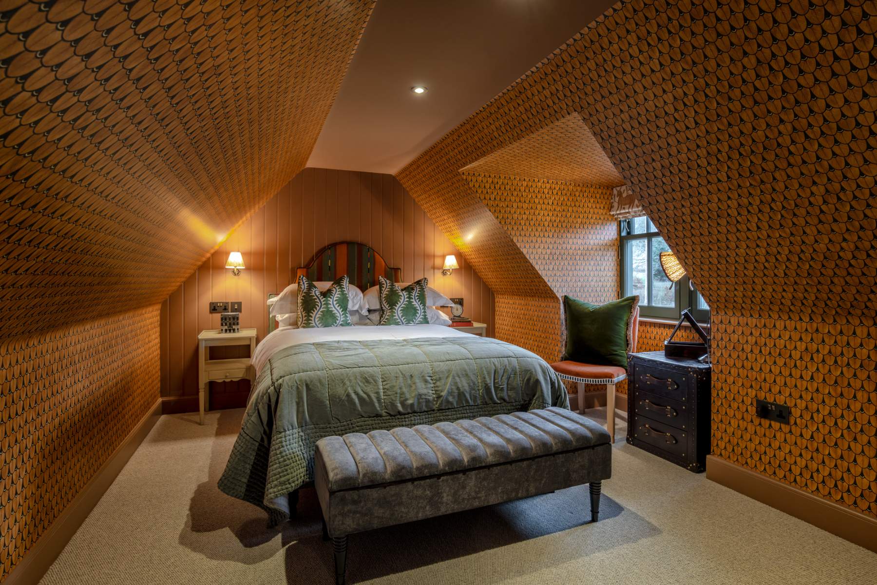 A bed with a sage green throw and patterned cushions sits in a mustard-papered attic room, a round leather stool and chest of drawers to the side.