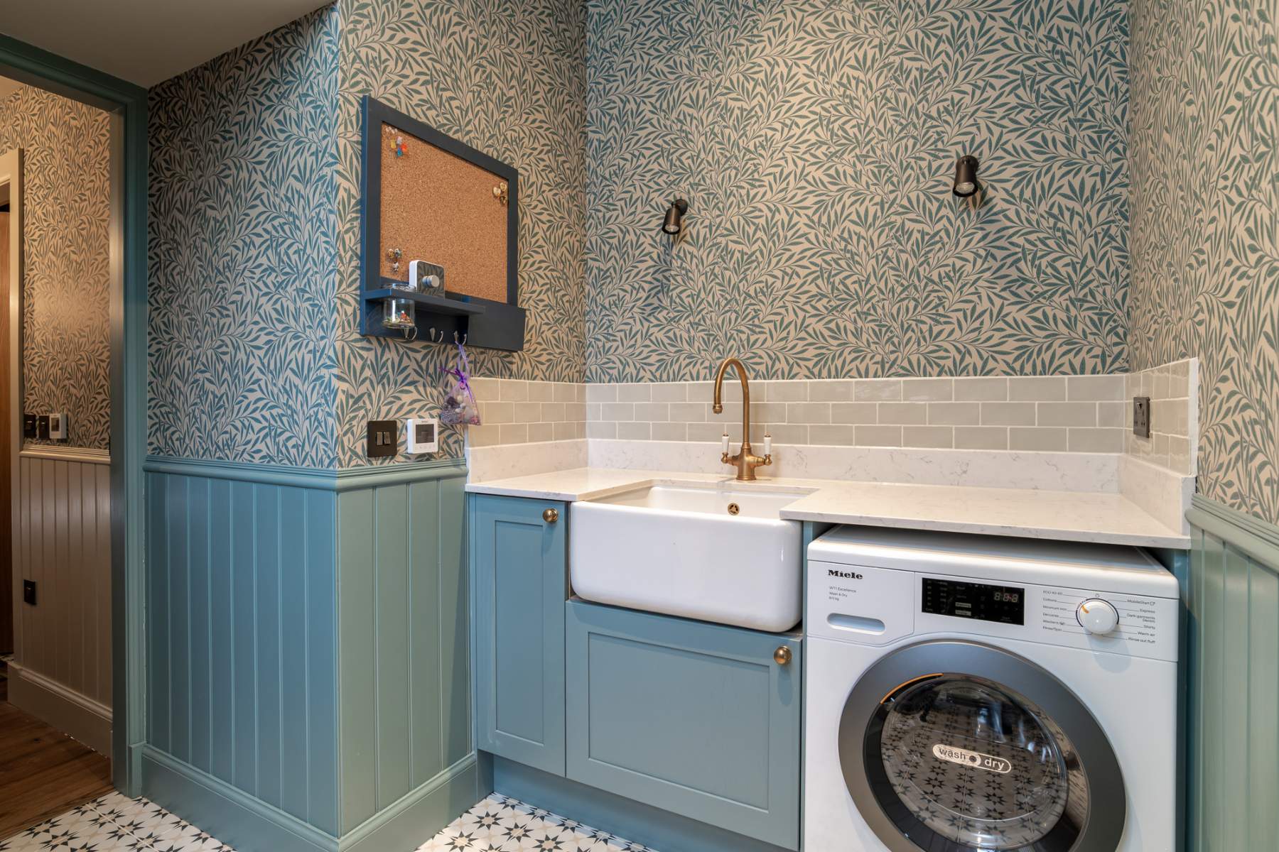A freestanding basin with a wall-mounted brass tap stands beside a washing machine, willow-patterned wallpaper above pale blue panelling.
