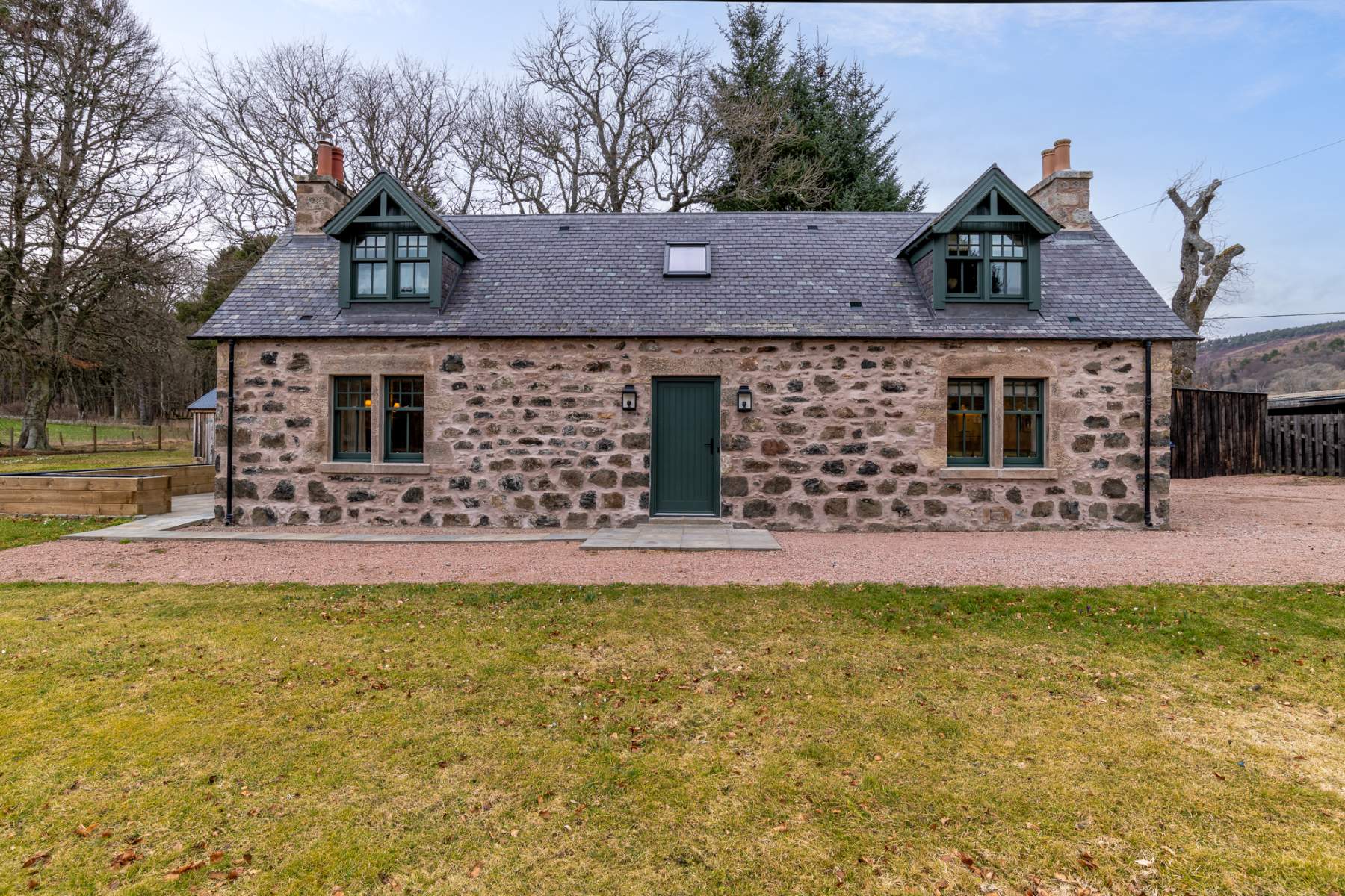 A single-storey stone cottage with matching gabled dormers and a green door stands behind a gravel drive.