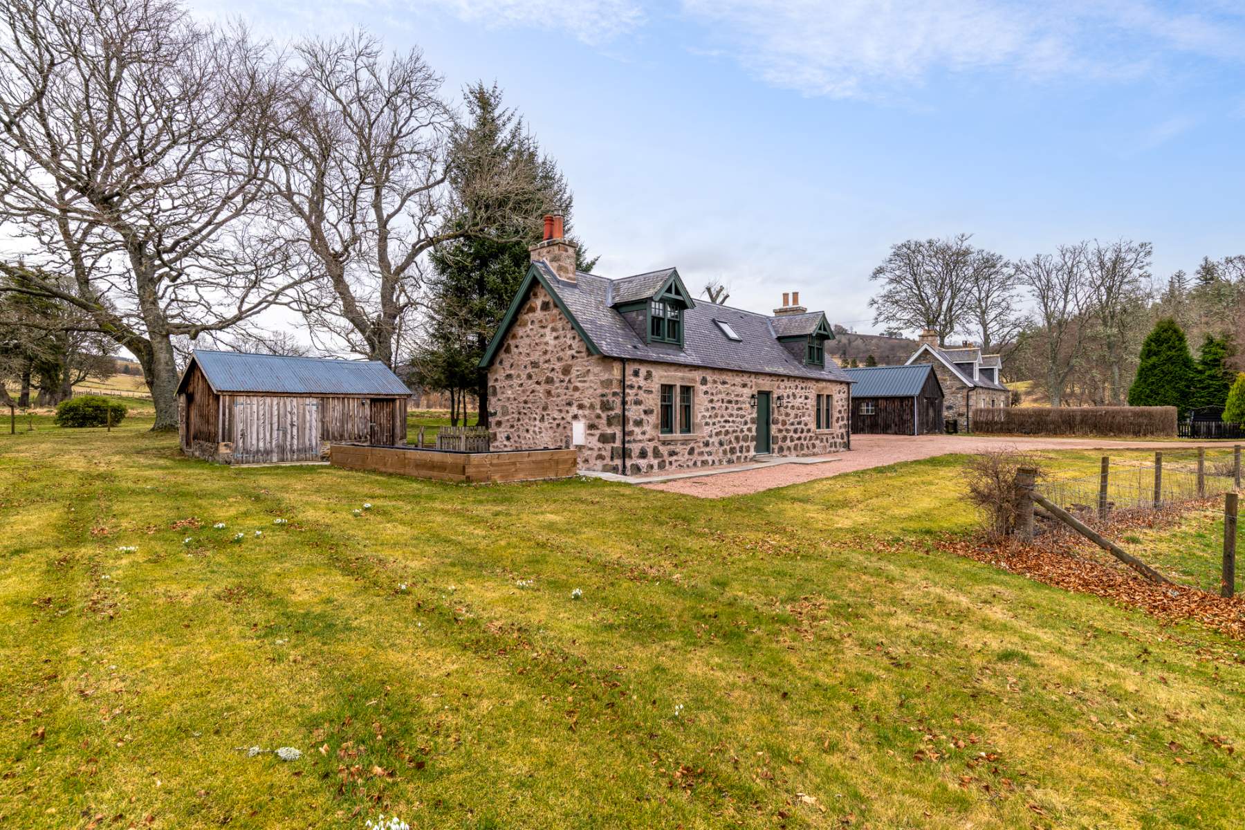 A cluster of stone farm buildings with green-framed windows sits among mature trees, a gravel path connecting them across the lawn.