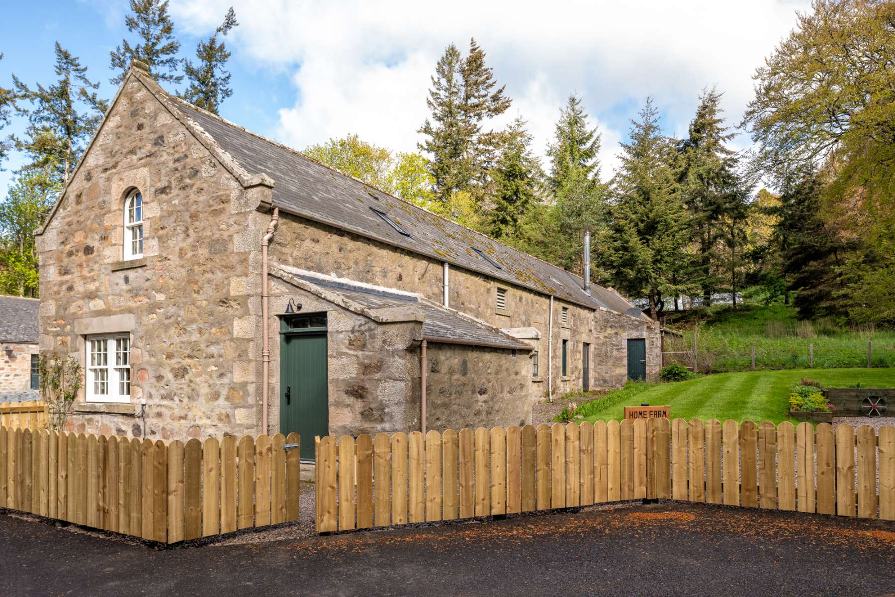 A long stone steading with a slated roof and green doors sits behind a timber picket fence, pine trees rising behind.