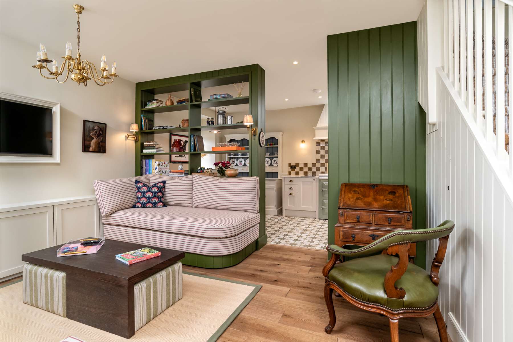 A striped daybed sits before green shelving of books and ornaments, a green leather armchair and antique writing bureau completing the room.