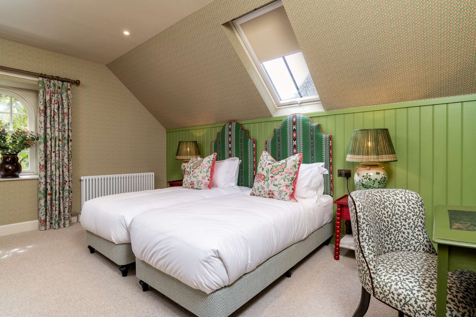 Twin beds with green striped headboards and floral cushions sit against green panelling, lamps lit on the shared bedside table.