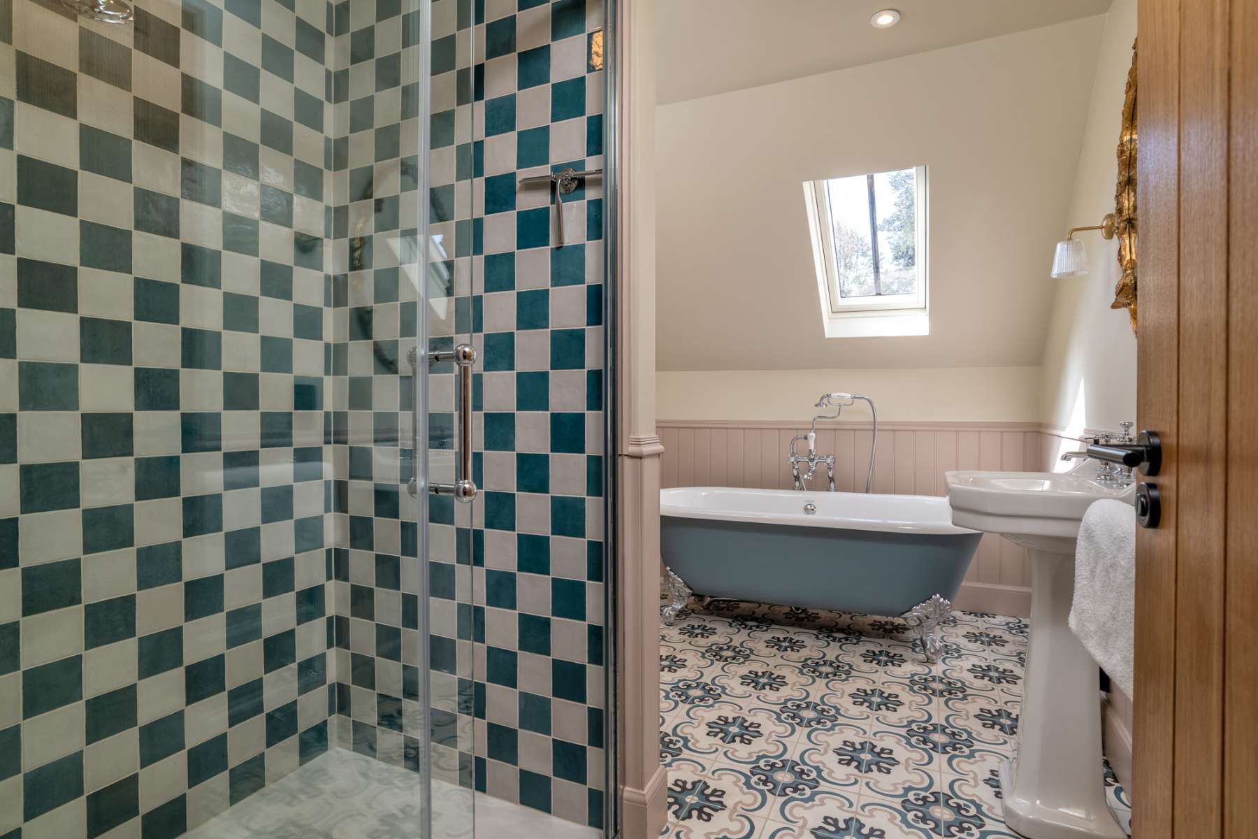 A glass shower cubicle in blue and white checked tiles stands beside a roll-top bath below a roof window, a pedestal basin to the right.
