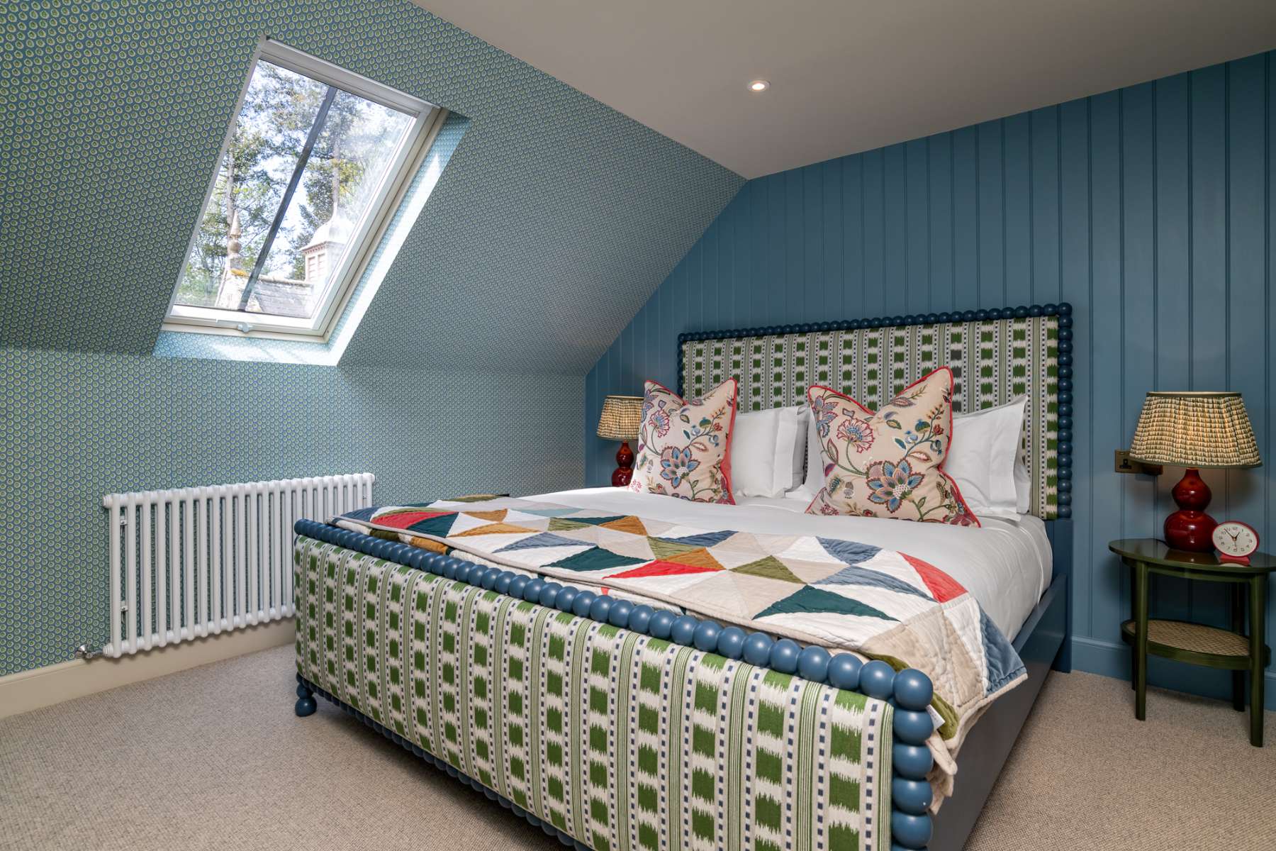 A patchwork-quilted bed with a green patterned headboard sits below a roof window, a red lamp lit on the bedside table.