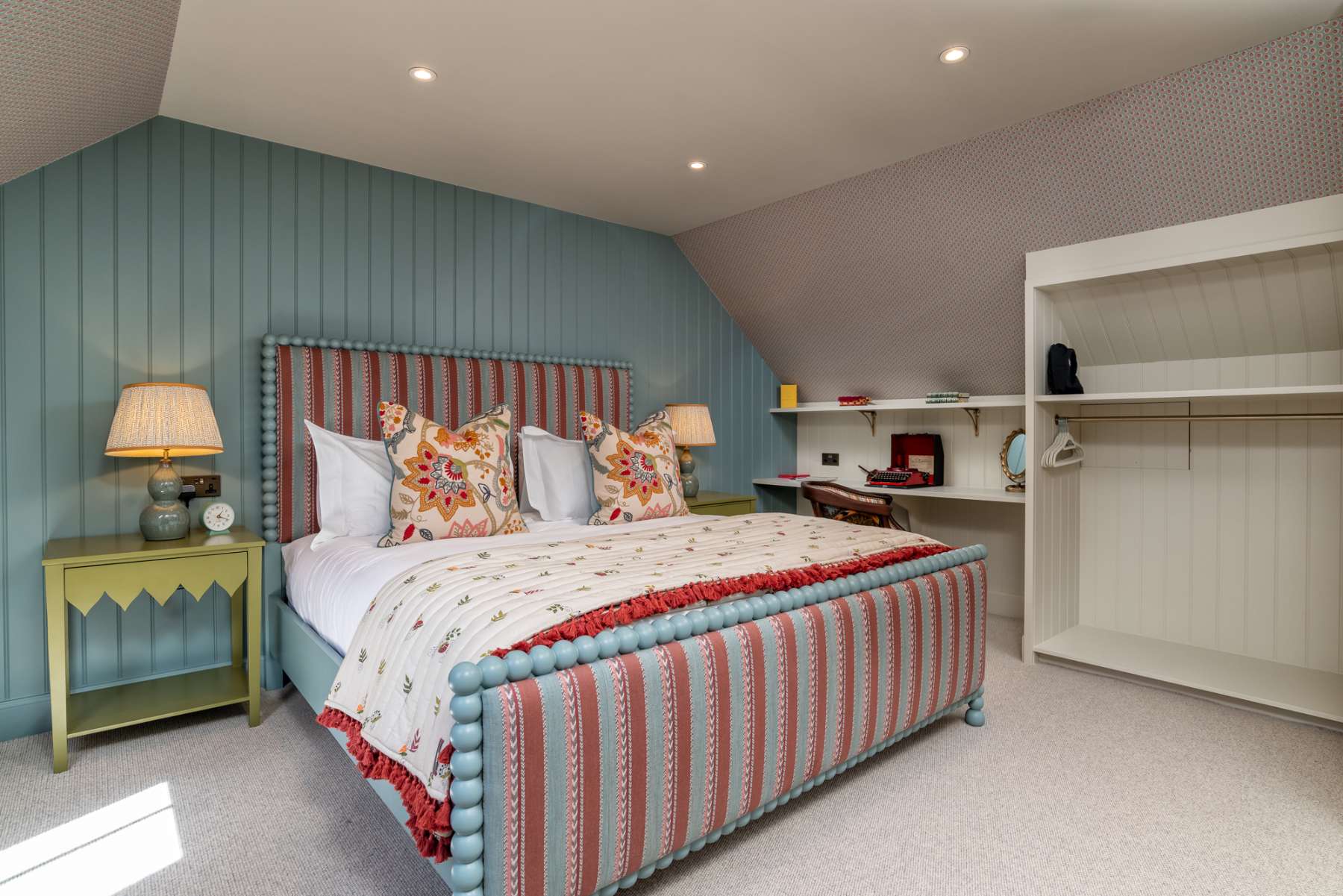 A bed with a blue striped upholstered frame and floral cushions sits against blue panelling, lamps lit on the tables either side.
