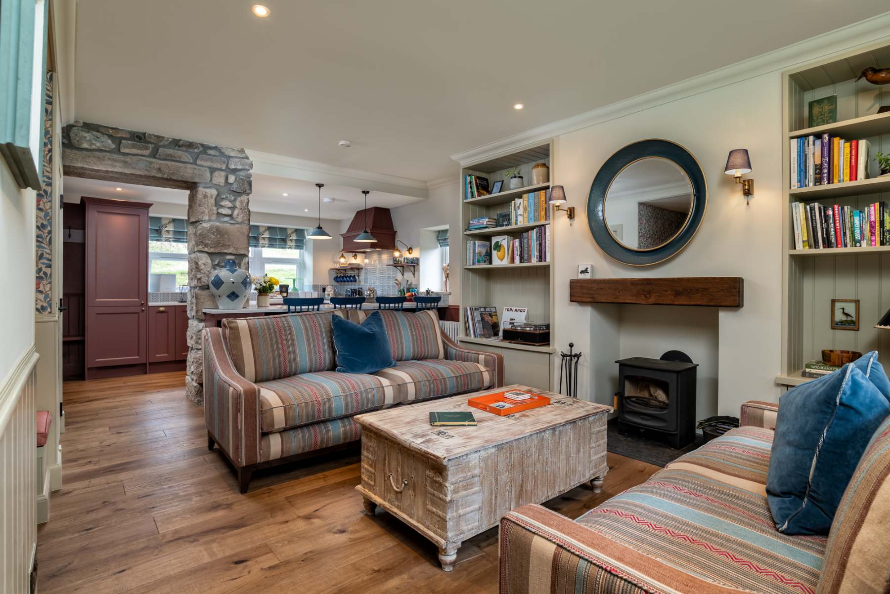 A stone archway opens from striped sofas to a kitchen beyond, a round mirror hanging above a wood-burning stove between bookshelves.