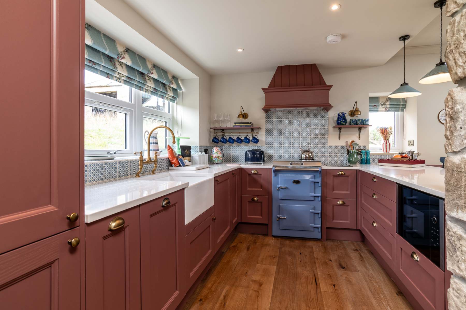 A blue range cooker stands beneath a dusty pink extractor hood in a kitchen with matching cabinetry and a patterned tile splashback.