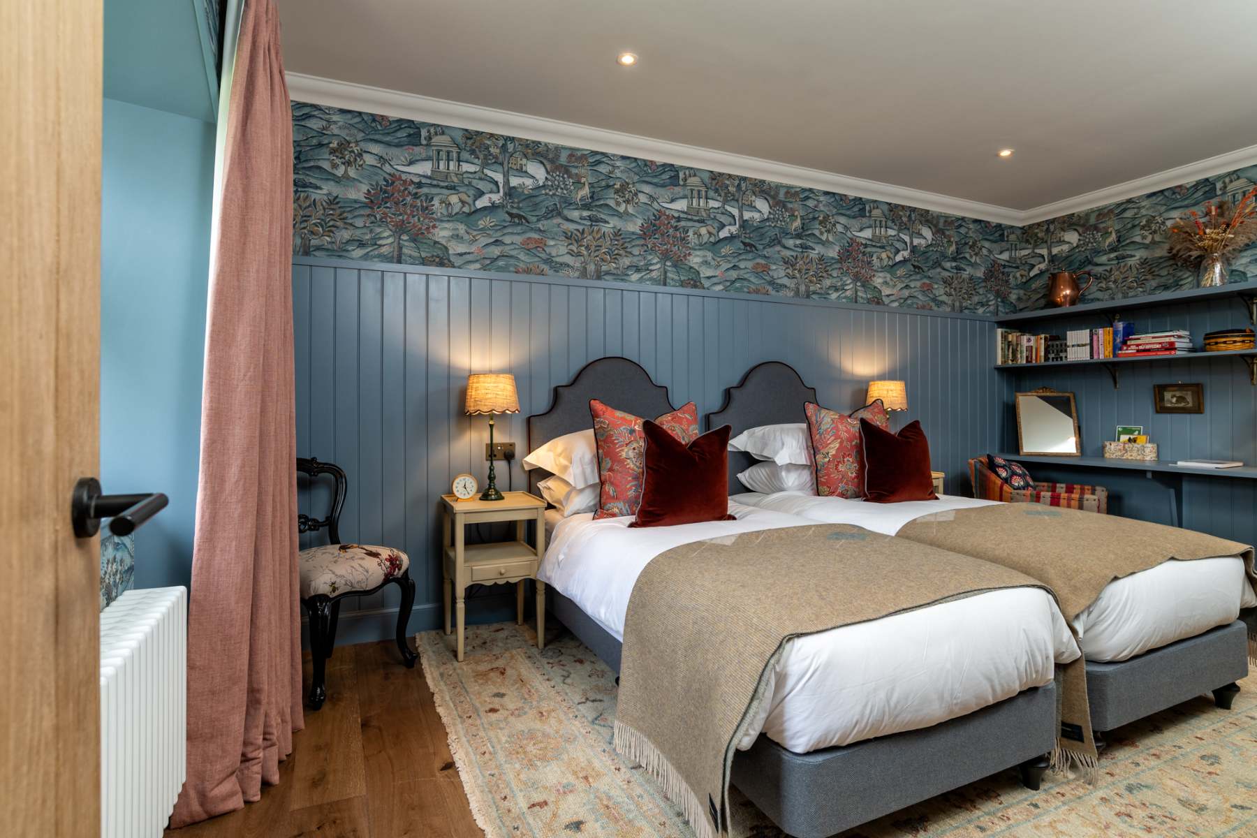 Twin beds with burgundy cushions sit beneath a bird-patterned wallpaper border, lamps lit on the bedside table and an antique chair by the pink curtain.