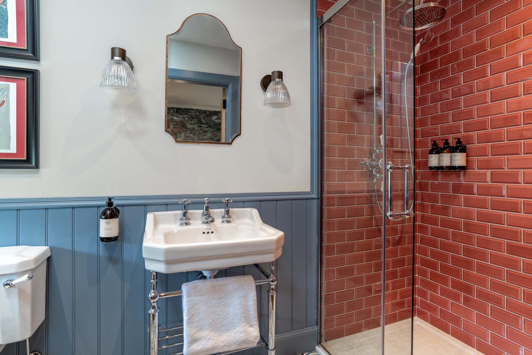An arched mirror hangs between wall sconces above a pedestal sink, red brick-effect tiles lining the shower beside it.