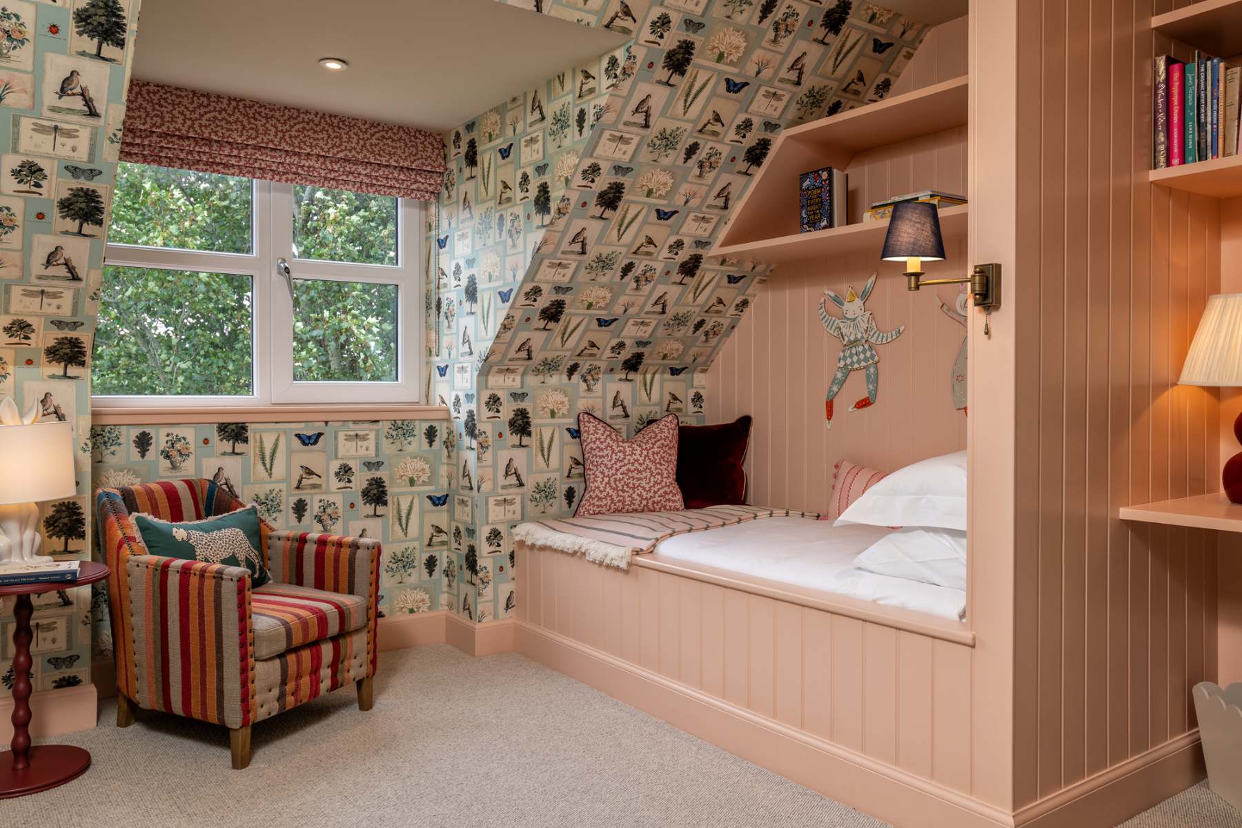 A striped armchair sits beside a bed alcove lined with nature-print wallpaper, a red cushion resting against the panelled headboard.
