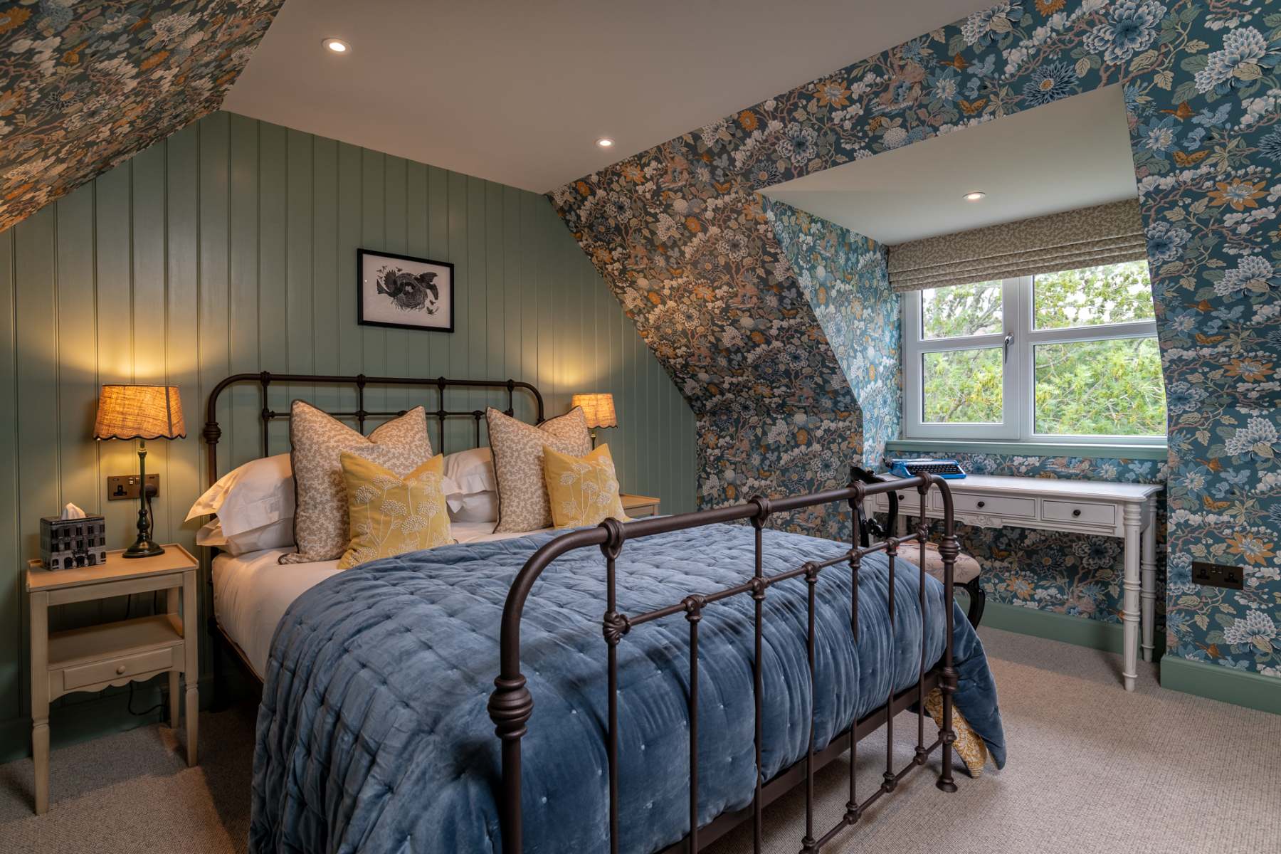 A dark iron bed frame with mustard cushions stands against sage green panelling, floral wallpaper on the sloped ceiling above a desk.