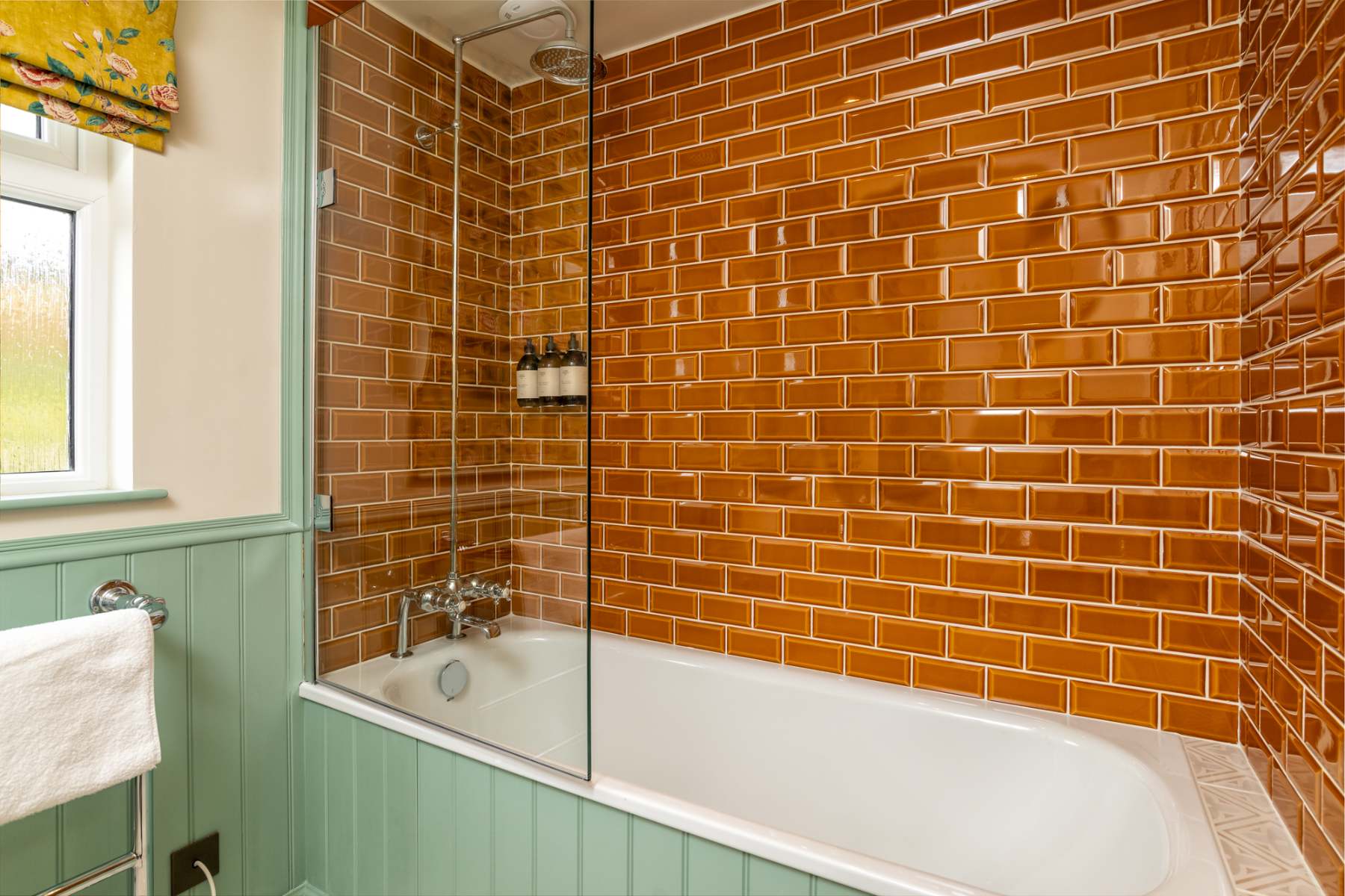 A glass screen shields a bath set against orange brick-effect tiles and green panelling, toiletry bottles on a small shelf.