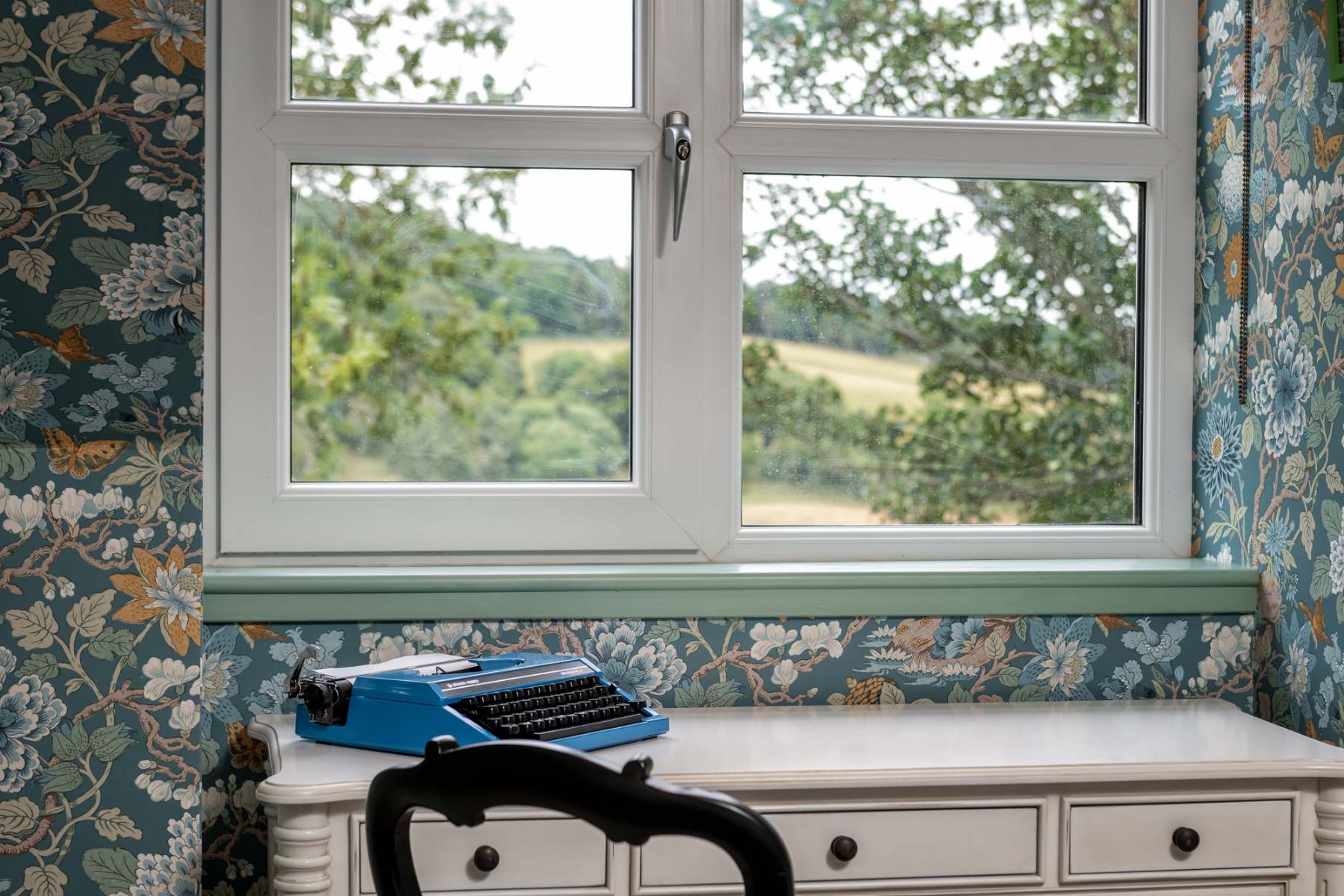 A vintage blue typewriter sits on a white dresser beneath a window looking out over fields and hills, floral wallpaper on the walls.