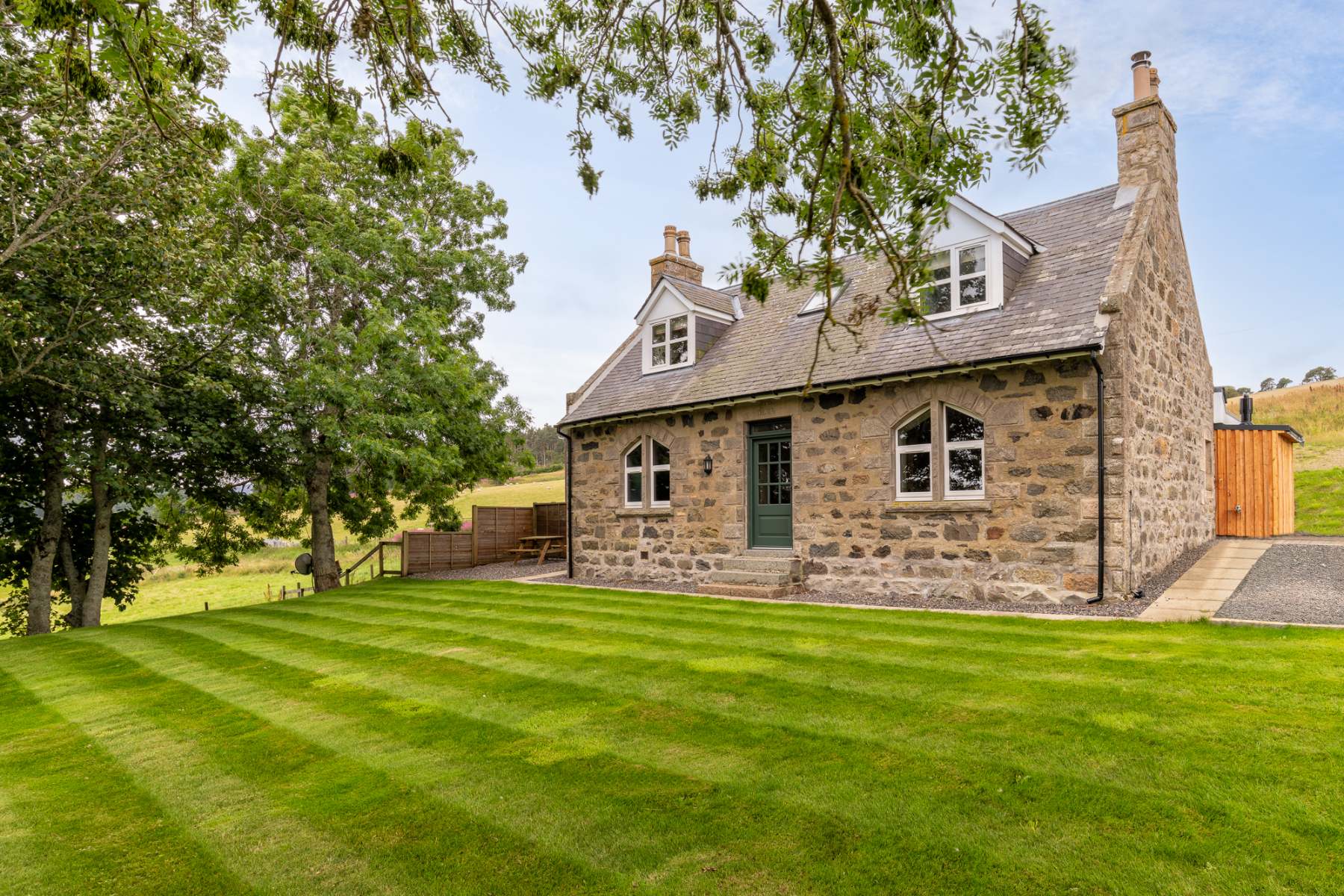 A stone farmhouse with dormer windows stands beyond a manicured lawn shaded by trees.