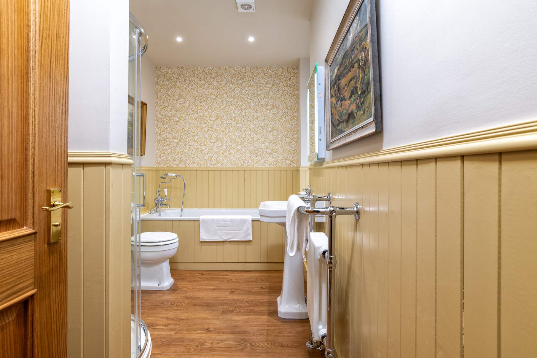 A bath with a folded towel sits below a framed painting, a toilet and heated towel rail to the left in a room papered with a small floral print.
