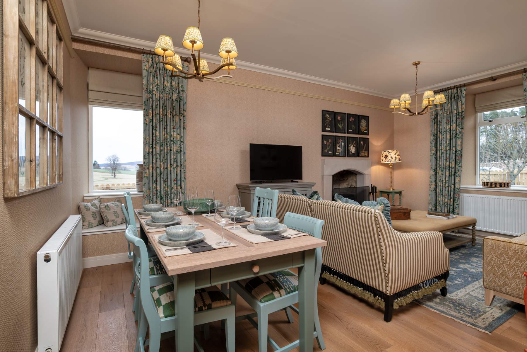 A dining table set with blue chairs stands before a sofa and fireplace, a mounted television on the wall behind.