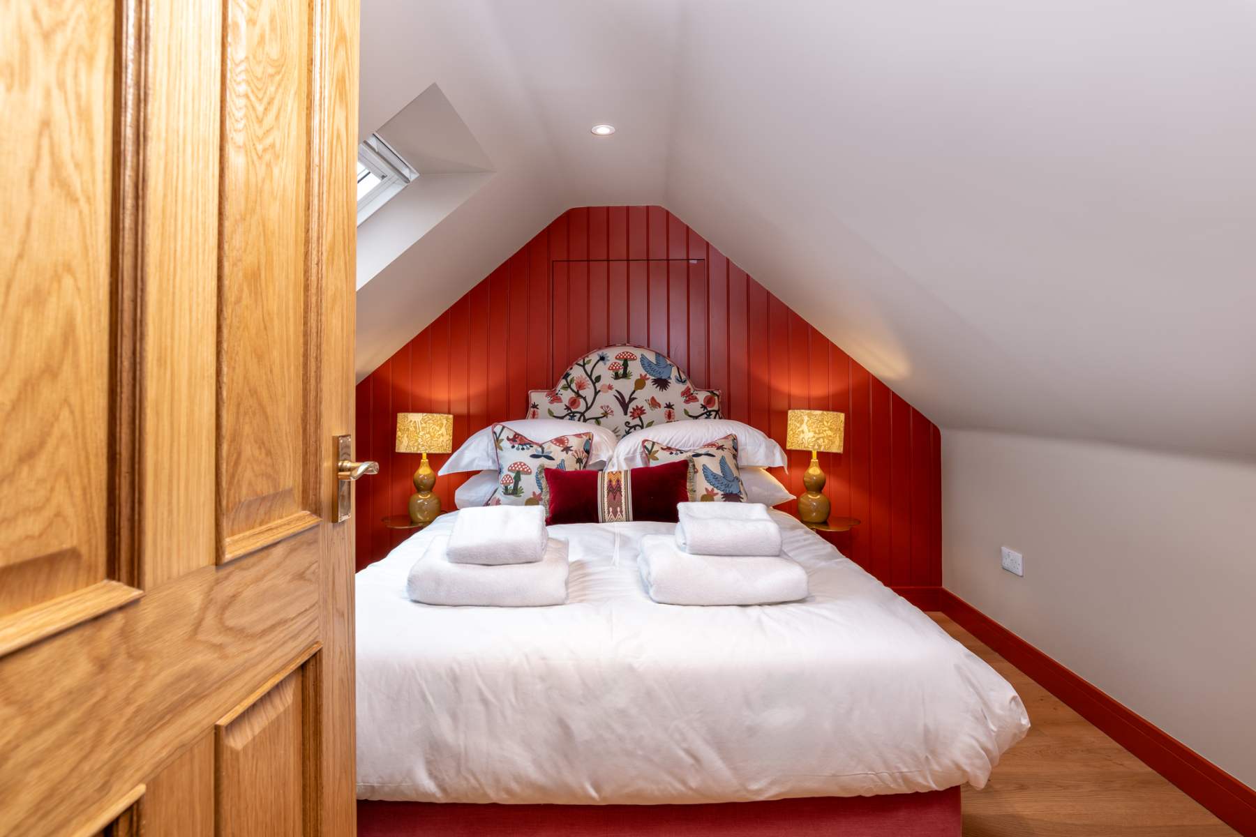 An oak panelled door stands open onto a red-walled attic bedroom, twin lamps lit either side of a floral headboard.