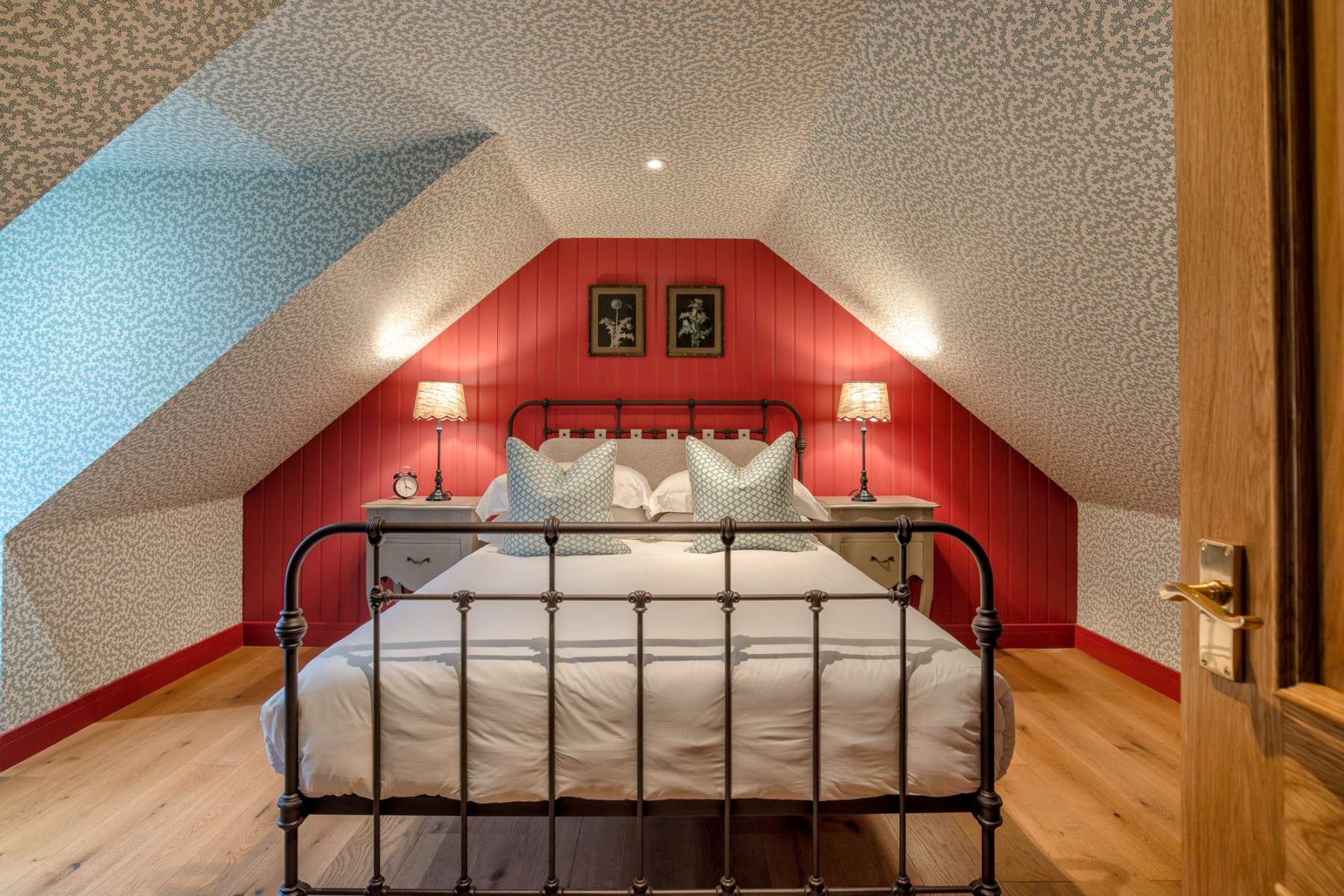 An iron bed with patterned cushions sits before a red panelled wall, lit lamps on bedside cabinets either side.