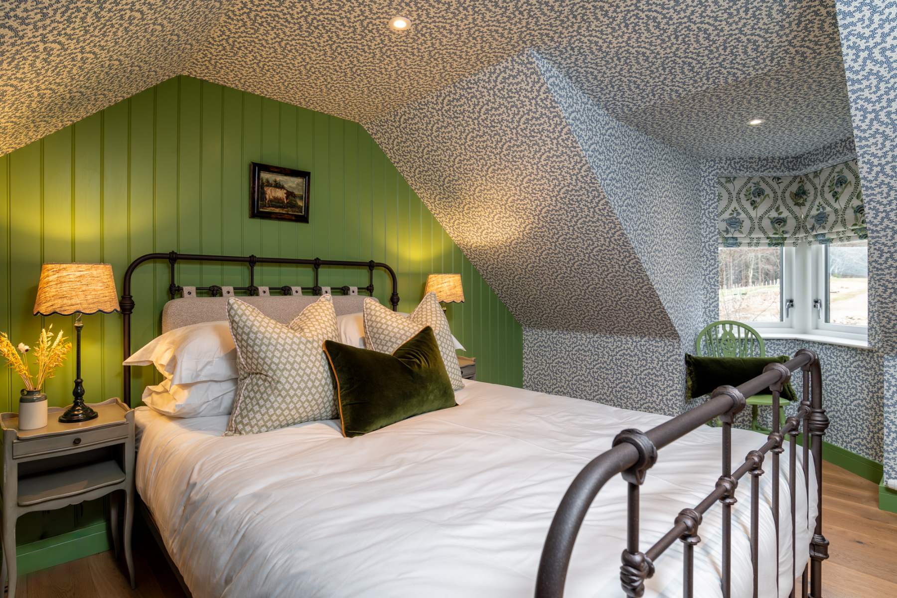 An iron bed with a green velvet cushion sits before dark green panelling, twin lamps lit on the bedside table and a window seat visible in the corner.