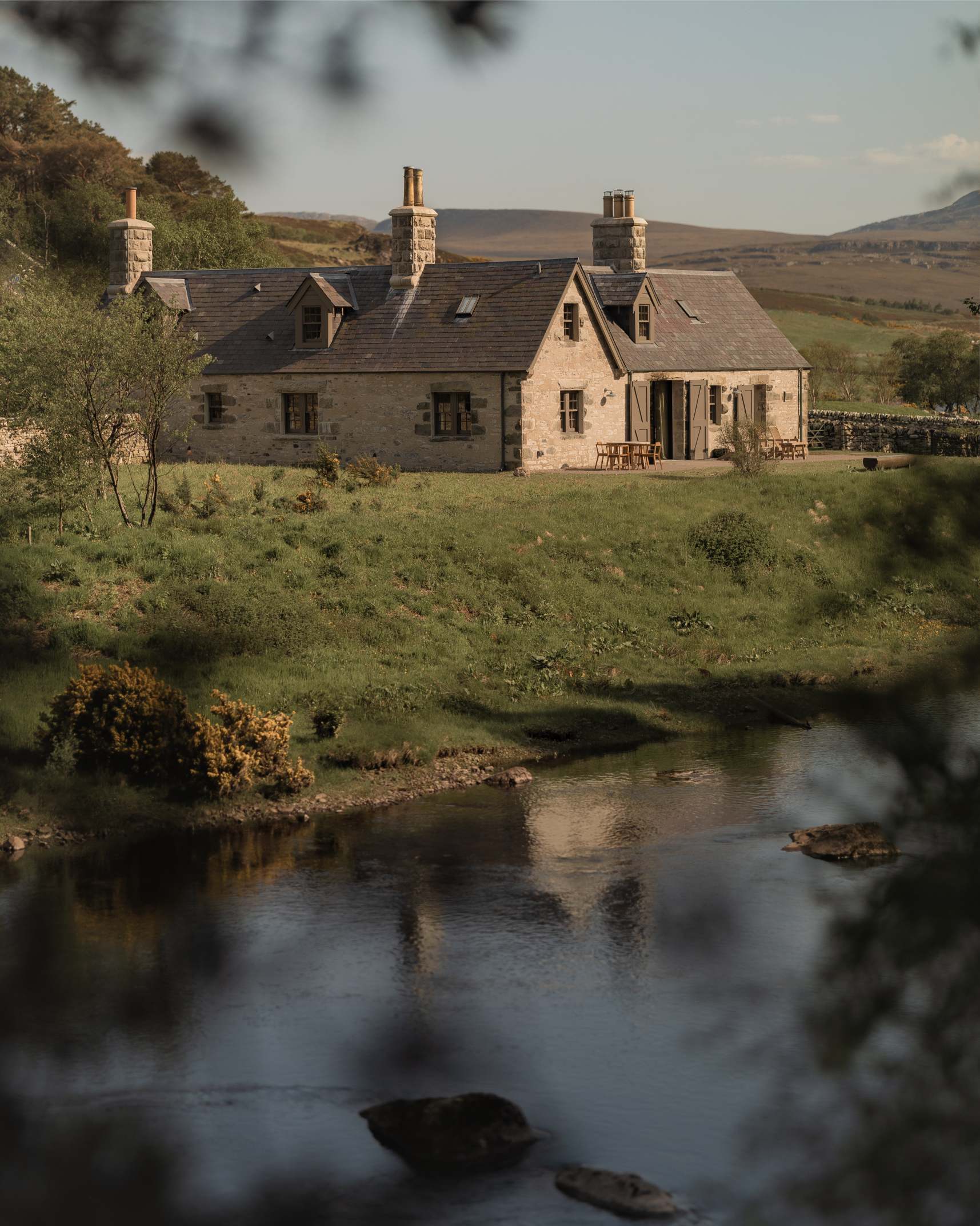 A stone farmhouse with multiple chimneys sits on a grassy bank above a river, hills rising behind, seen through overhanging leaves.