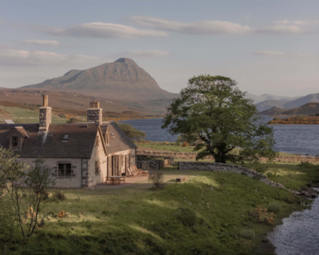 A stone cottage with two chimneys sits above a loch, a mountain rising behind and a dry stone wall enclosing the lawn.