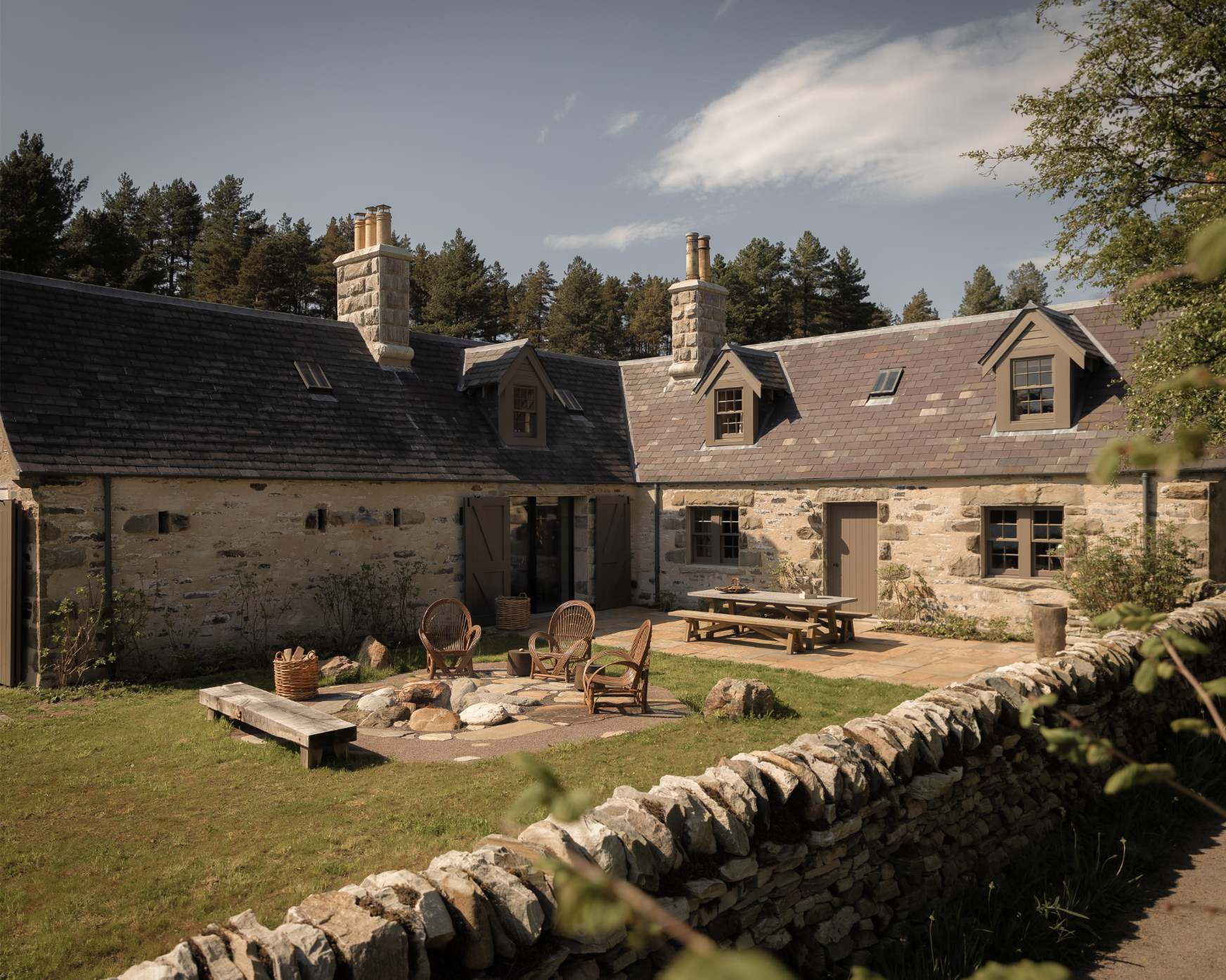 A stone house with multiple chimneys and dormer windows sits behind a courtyard set with woven chairs and a picnic bench, pine trees rising behind.
