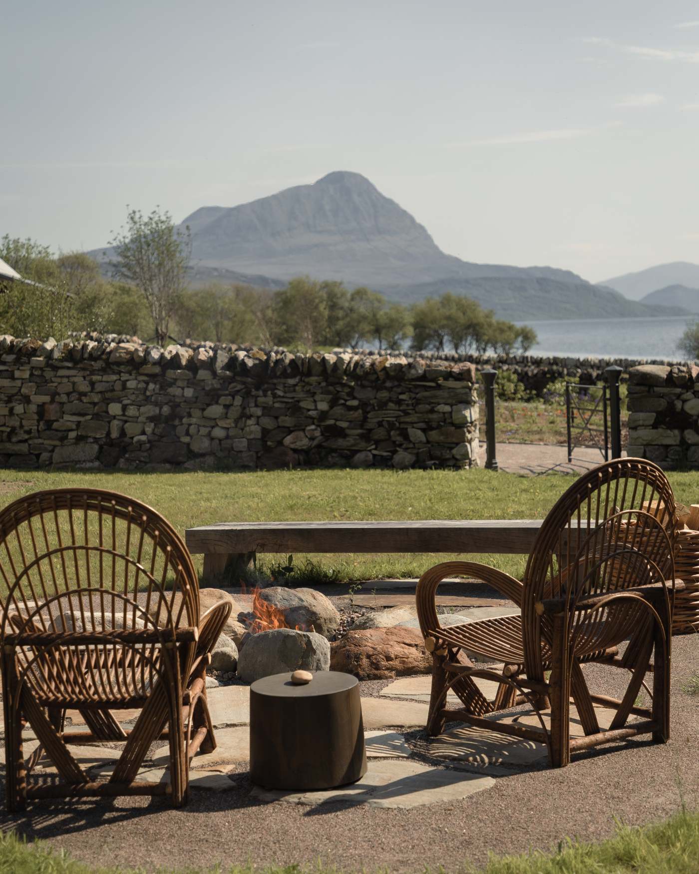 Woven chairs and a bench surround a fire pit on a patio, a dry stone wall framing a view of the loch and mountain beyond.