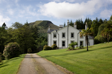Glenstriven House, a white Victorian house with grey stone corners, at the top of a gravel drive between lawns, with a wooded hill behind.