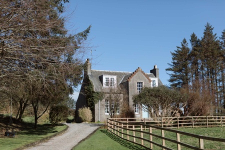 A grey stone house with crow-stepped gables sits behind a post and rail fence, pine trees flanking a gravel drive.