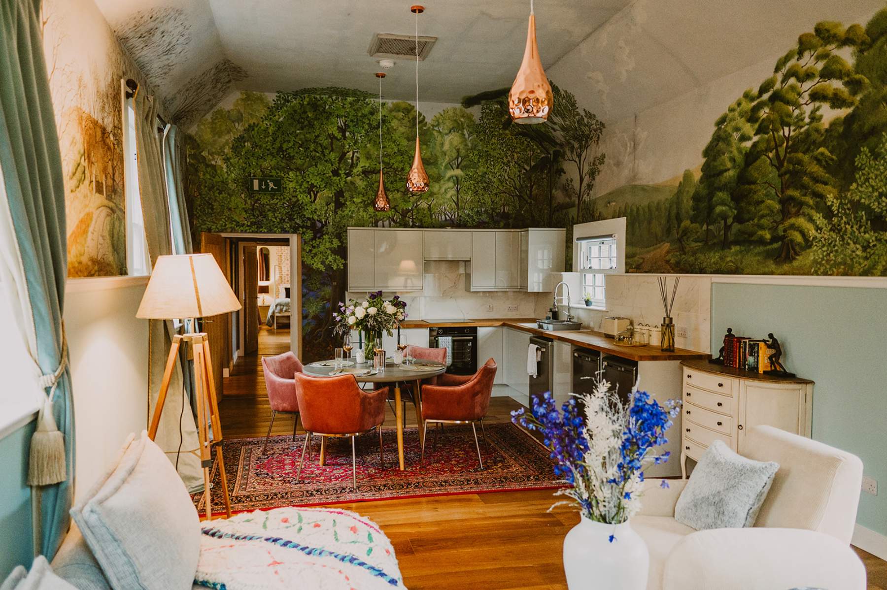 A pink dining table with rattan chairs stands before a fitted kitchen, walls painted with a woodland mural beneath pendant lights and cream sofas in the foreground.