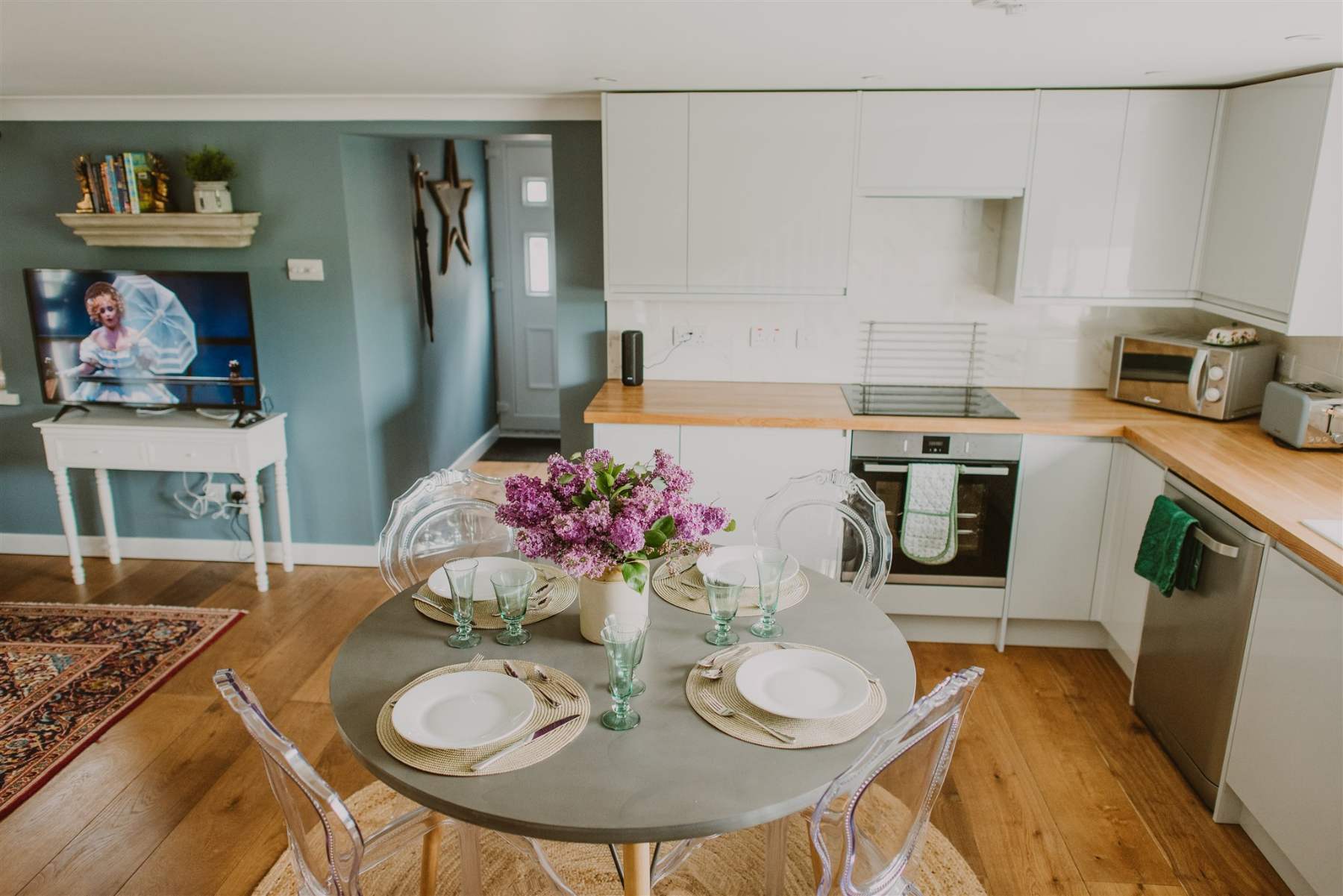 A round grey dining table set with glasses and a vase of flowers stands before a white kitchen, a mounted television and console table to the left.