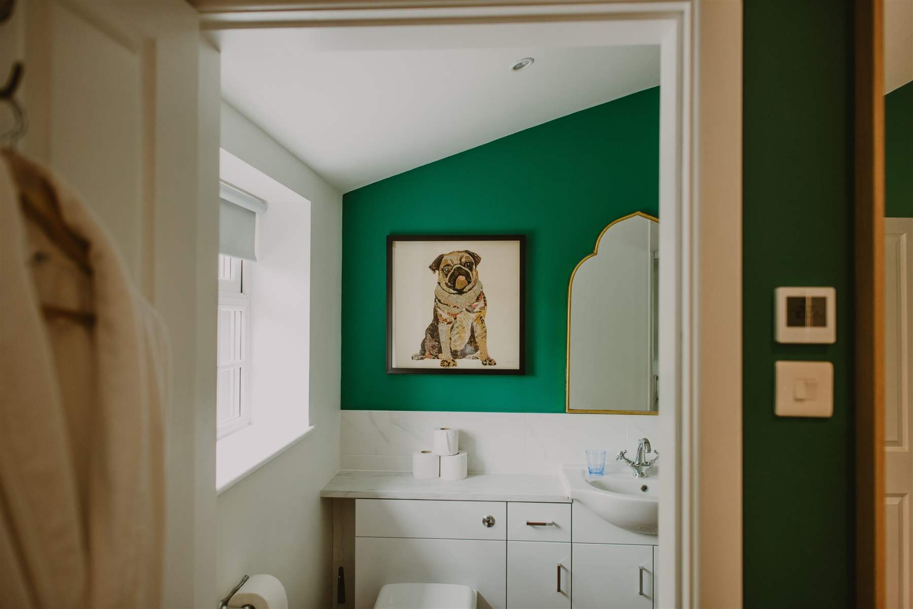 A framed illustration of a pug hangs on a green wall above a bathroom vanity, a toilet and roll of paper in the foreground.