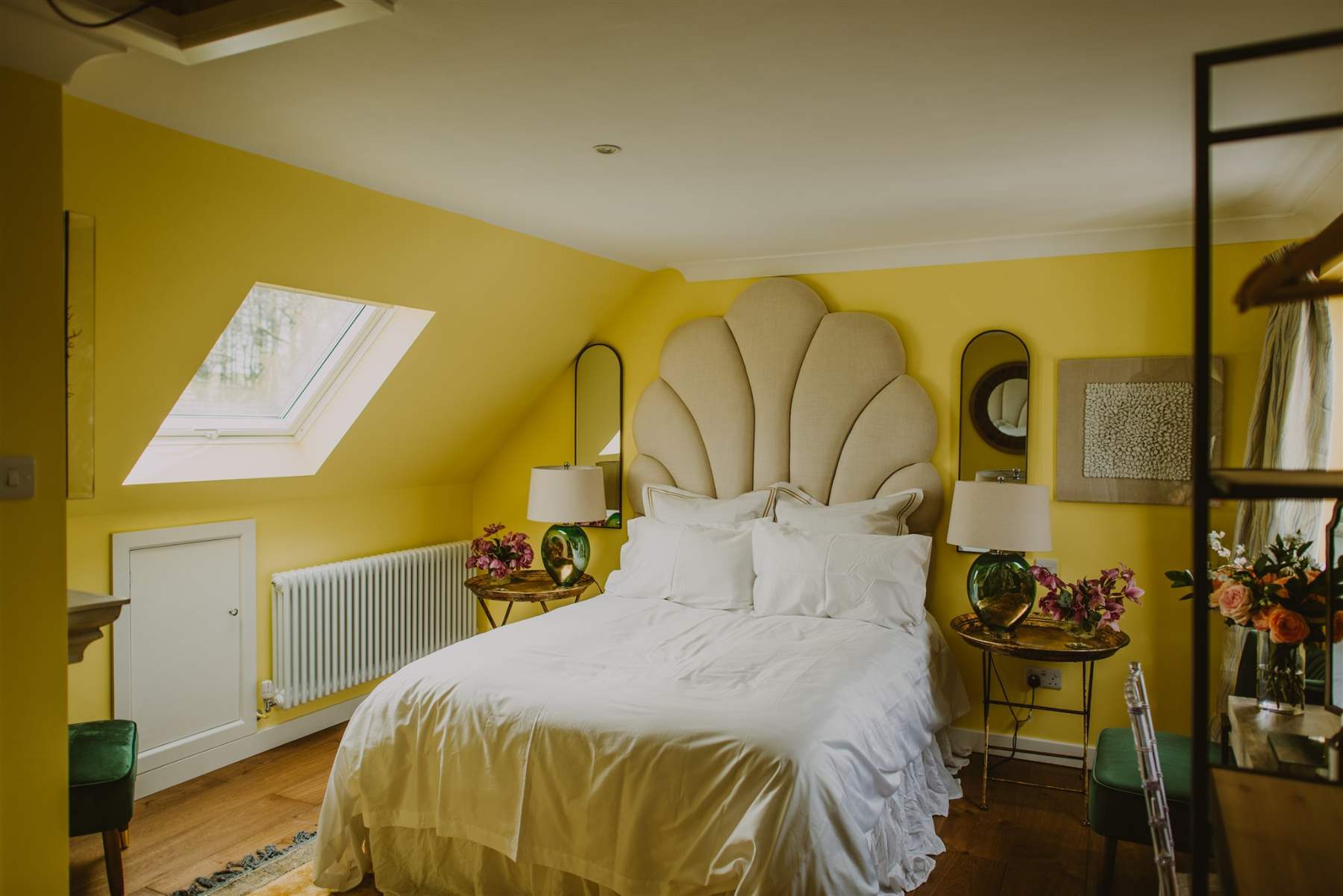 A shell-shaped upholstered headboard sits between two mirrors on a yellow wall, lamps and vases of flowers on the bedside tables either side.
