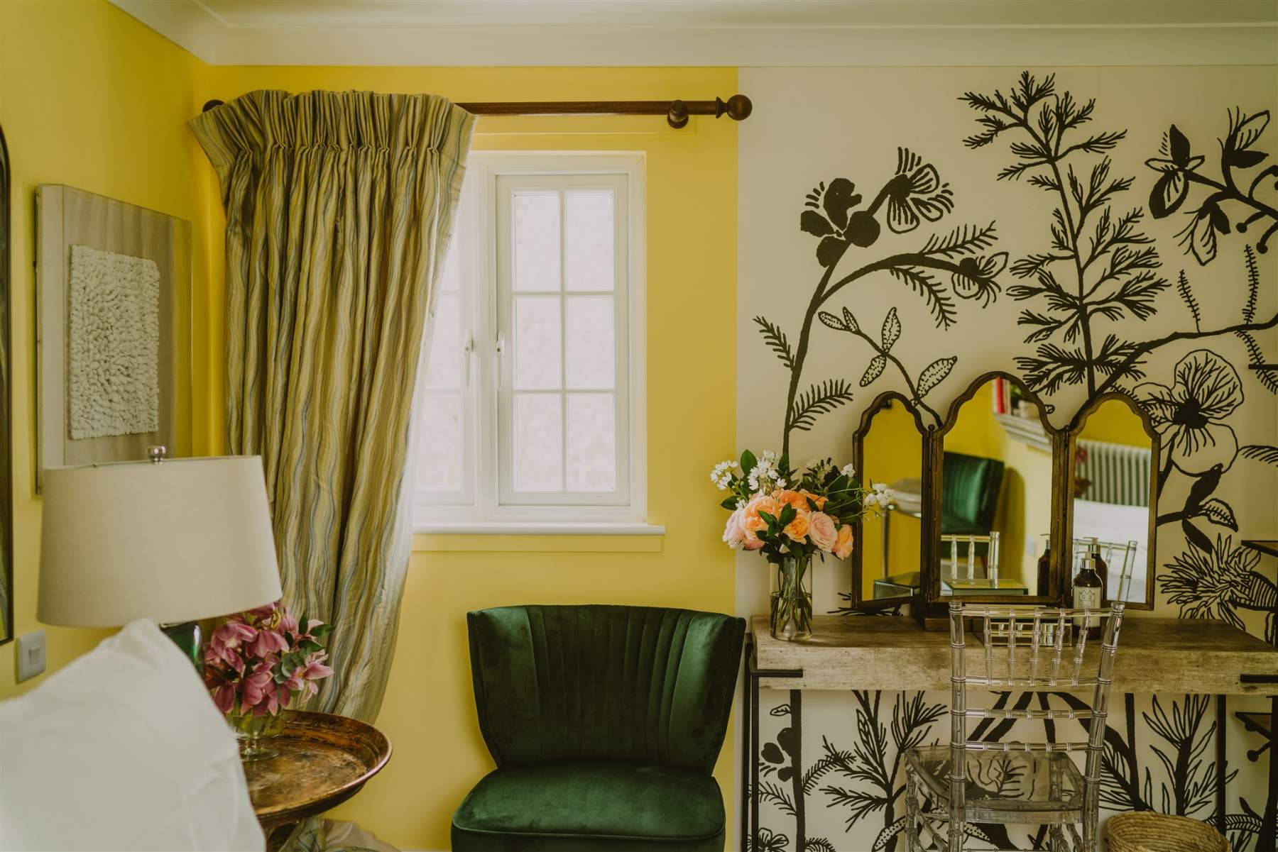 A green velvet chair sits before a dressing table with three mirrors, hand-painted botanical branches decorating the yellow wall beside striped curtains.