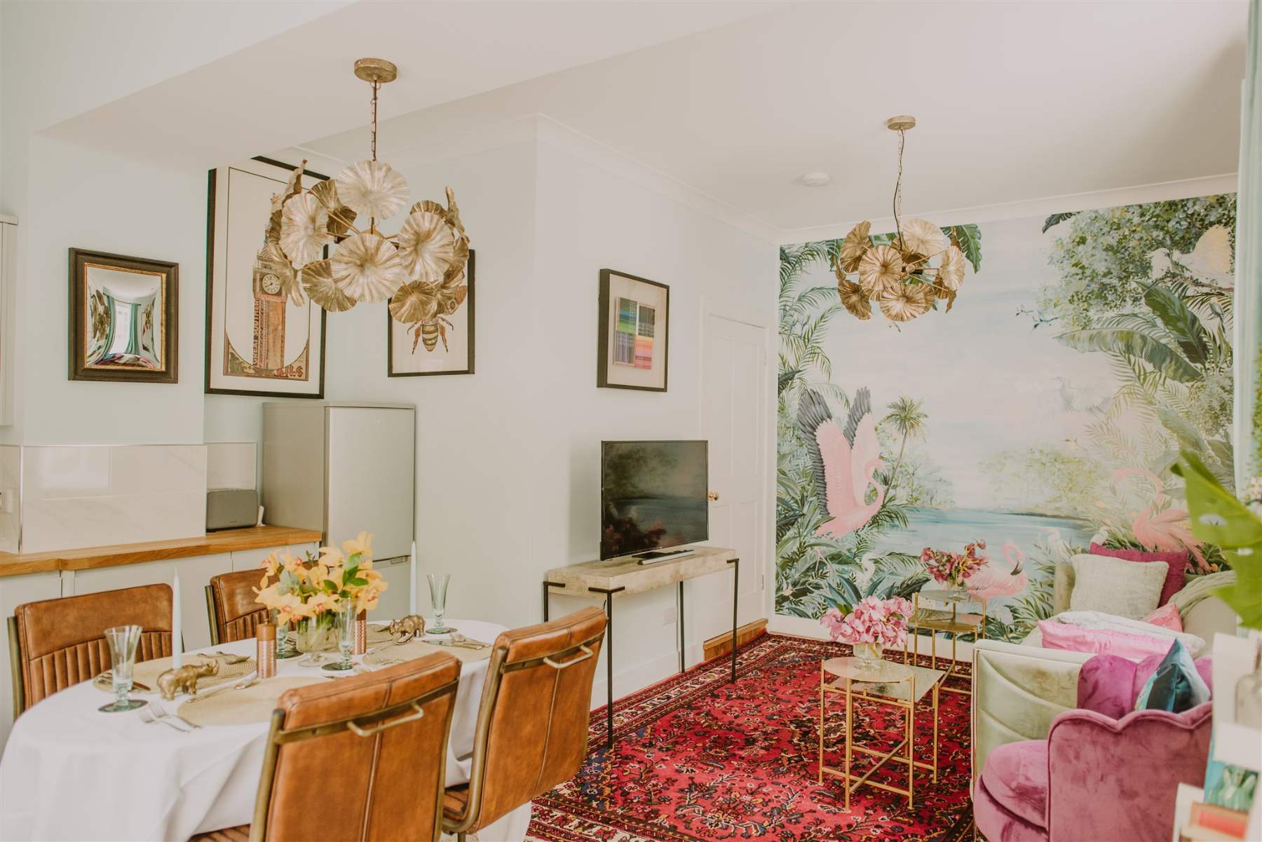 A dining area with patterned wallpaper of large leaves and flowers, seen past a framed mirror and prints on the near wall.