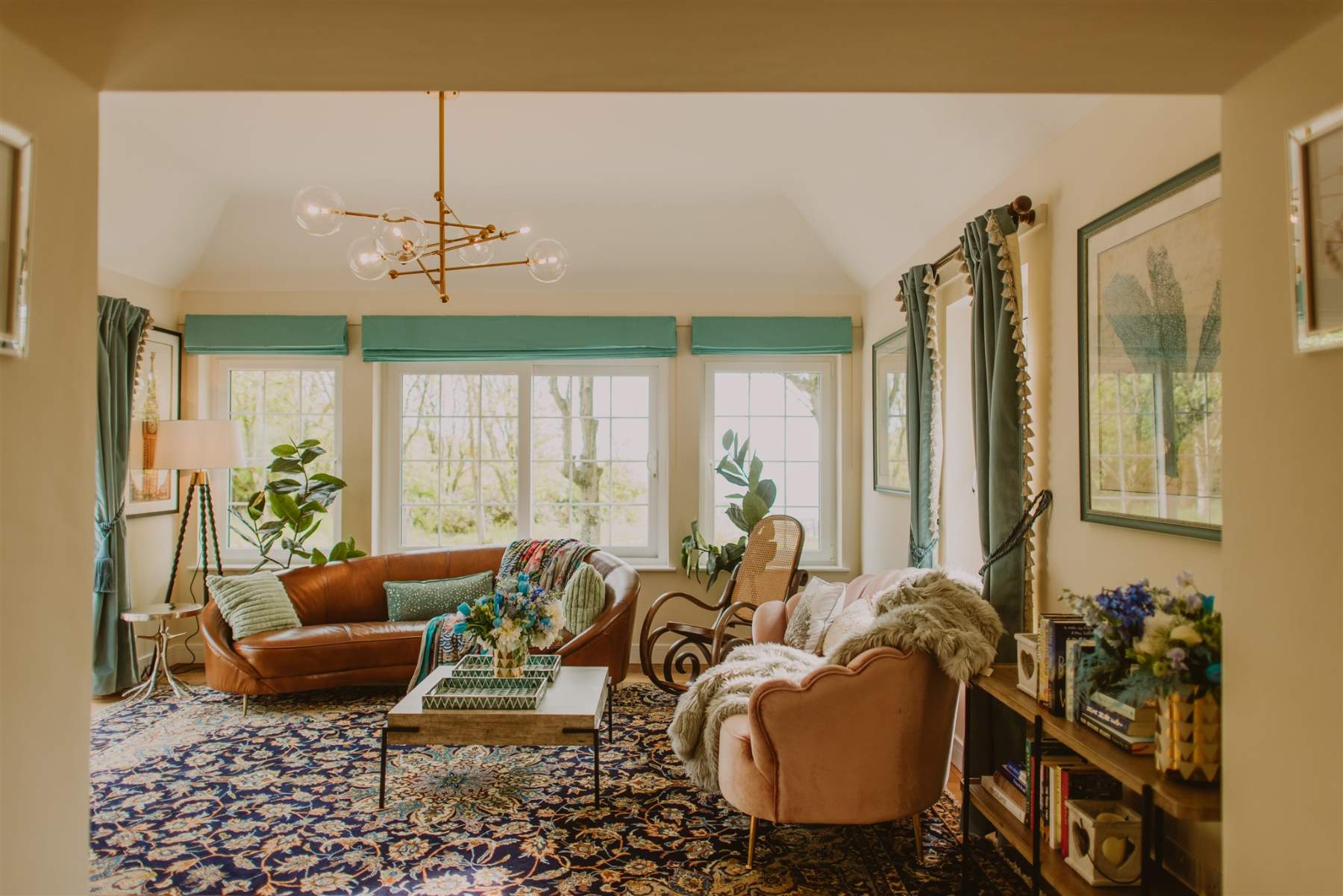 A rust leather sofa and pink armchairs surround a low table in a sitting room with teal roman blinds, a starburst pendant light above and shelves of books to the side.