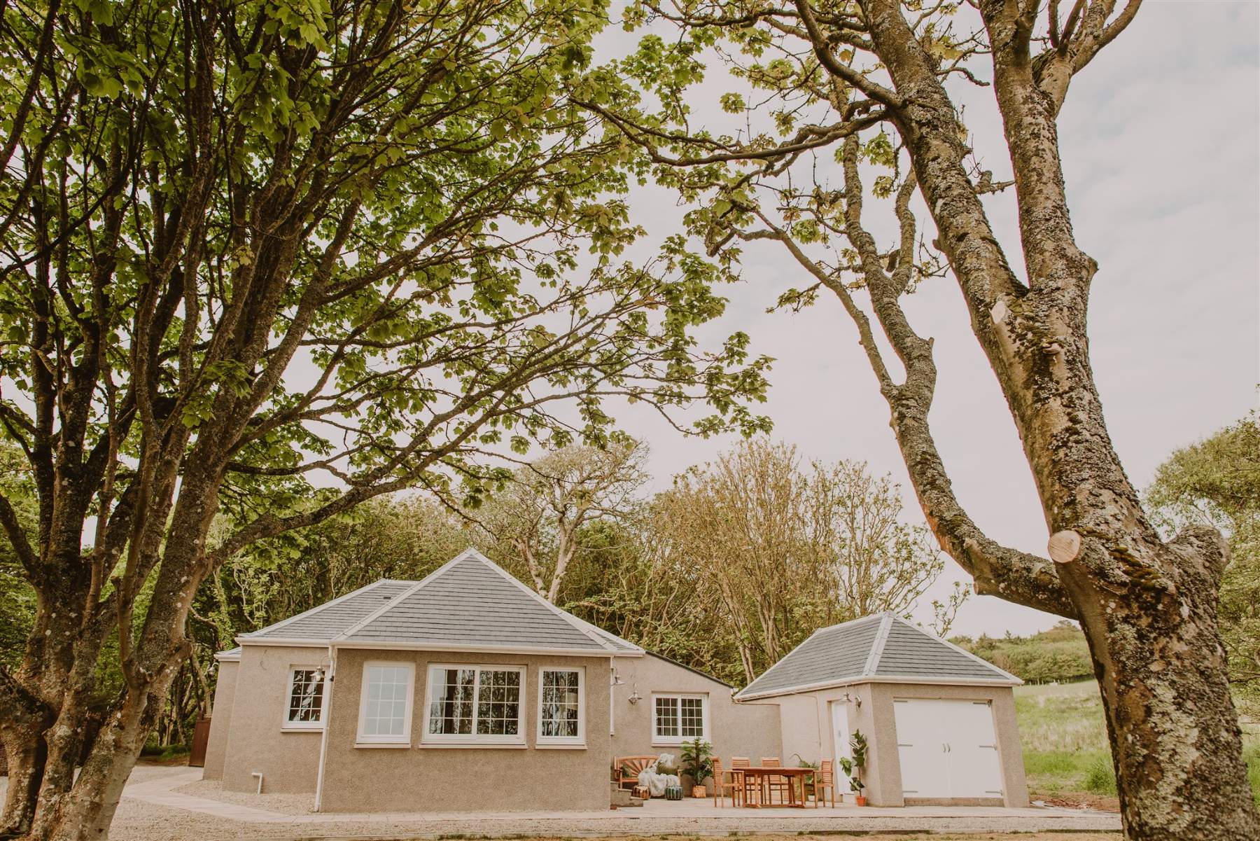 A pink harled house with a hipped roof sits behind a gravel drive, trees framing the building on either side.
