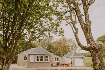 A pink harled house with a hipped roof sits behind a gravel drive, trees framing the building on either side.