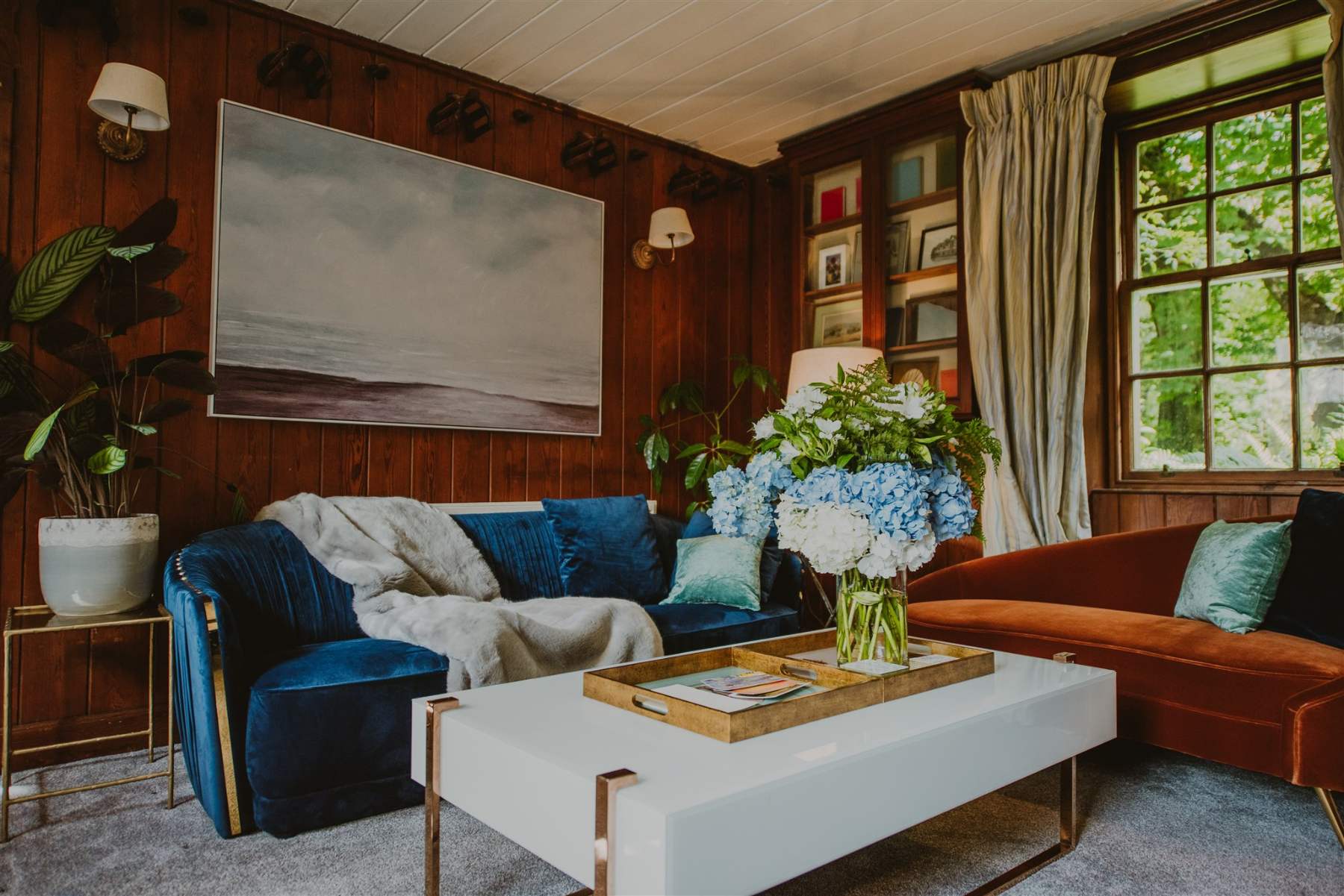 A navy velvet sofa and rust-coloured sofa flank a white coffee table set with hydrangeas, a framed seascape painting on the panelled wall above.