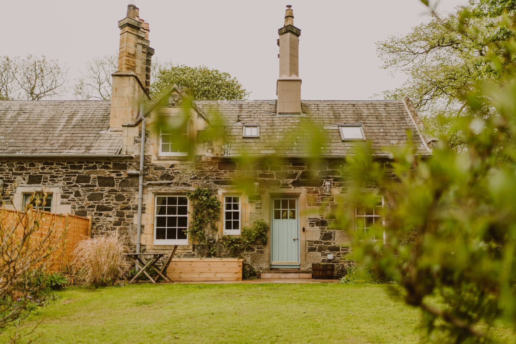 A stone cottage with two chimneys and a pale blue door sits behind a lawn, a bench and planter on the gravel path in front.