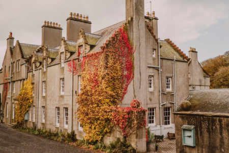 A stone country house facade is partly covered in red and gold autumn ivy climbing past its dormer windows and chimney stacks.